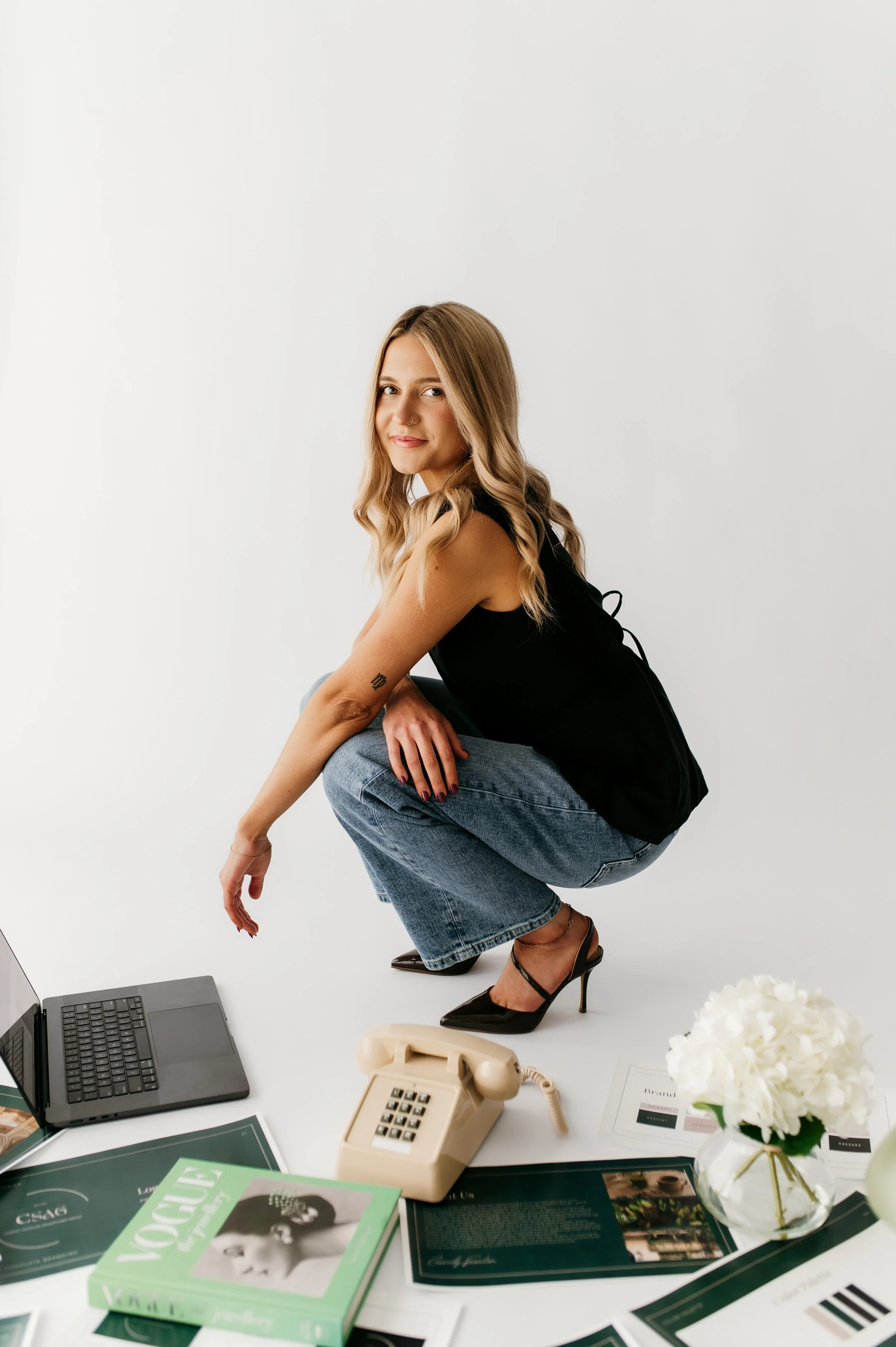 A woman with blonde hair crouching on a white office floor with a workspace in front of her that includes a laptop, a beige telephone, magazines, and a vase of white flowers. She is wearing a black sleeveless top, blue jeans, and black high-heeled shoes, and is smiling at the camera.
