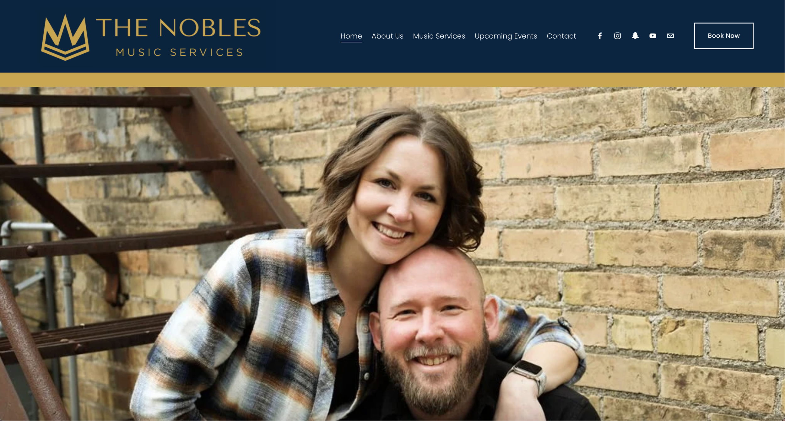 A smiling woman with brown hair wearing a plaid shirt and a man with a beard and short hair, also smiling, leaning against a brick wall outdoors.