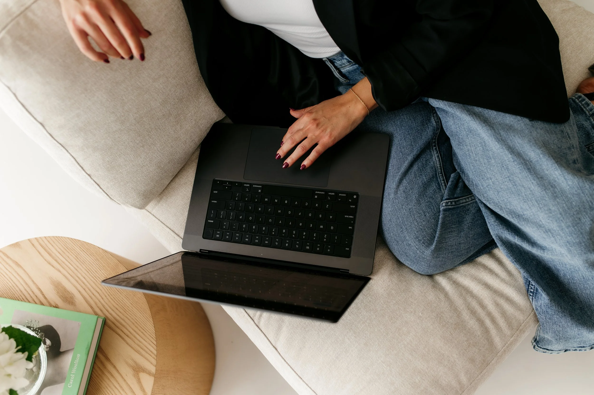 Person sitting on a beige sofa with a black laptop on their lap, wearing jeans and a blazer, in a cozy indoor setting.