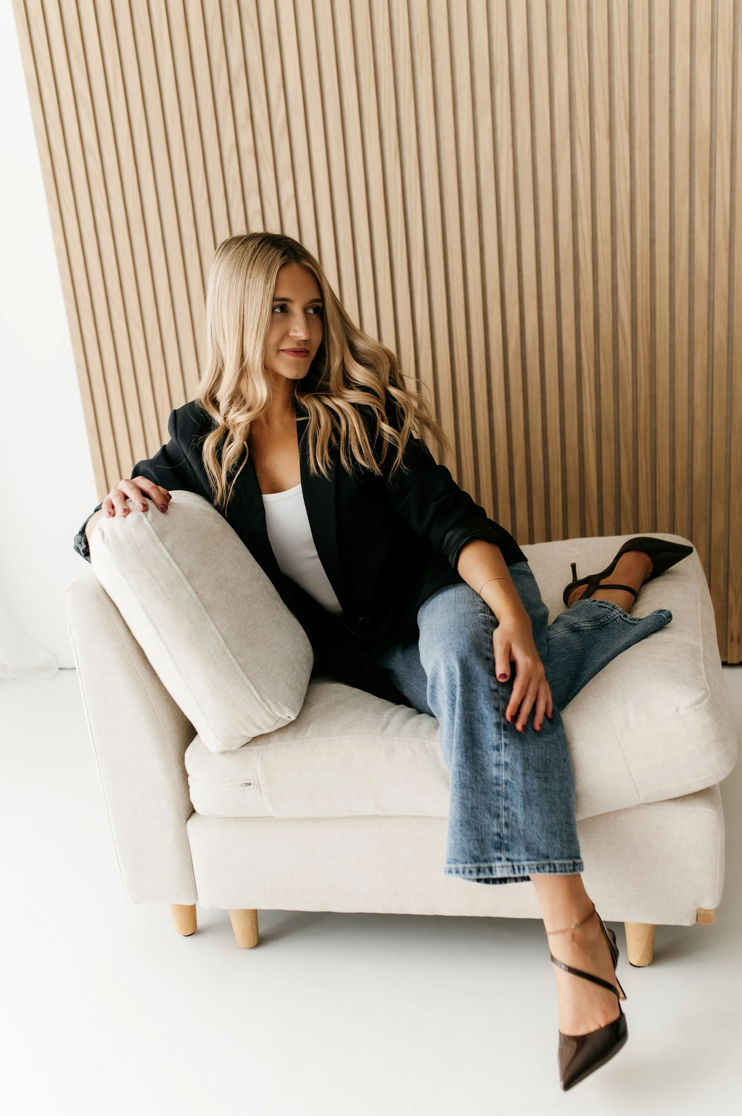 A woman with long, wavy blonde hair sitting on a beige armchair with wooden legs in a room with a wooden slatted wall, wearing a black blazer, white top, blue jeans, and black high heels.