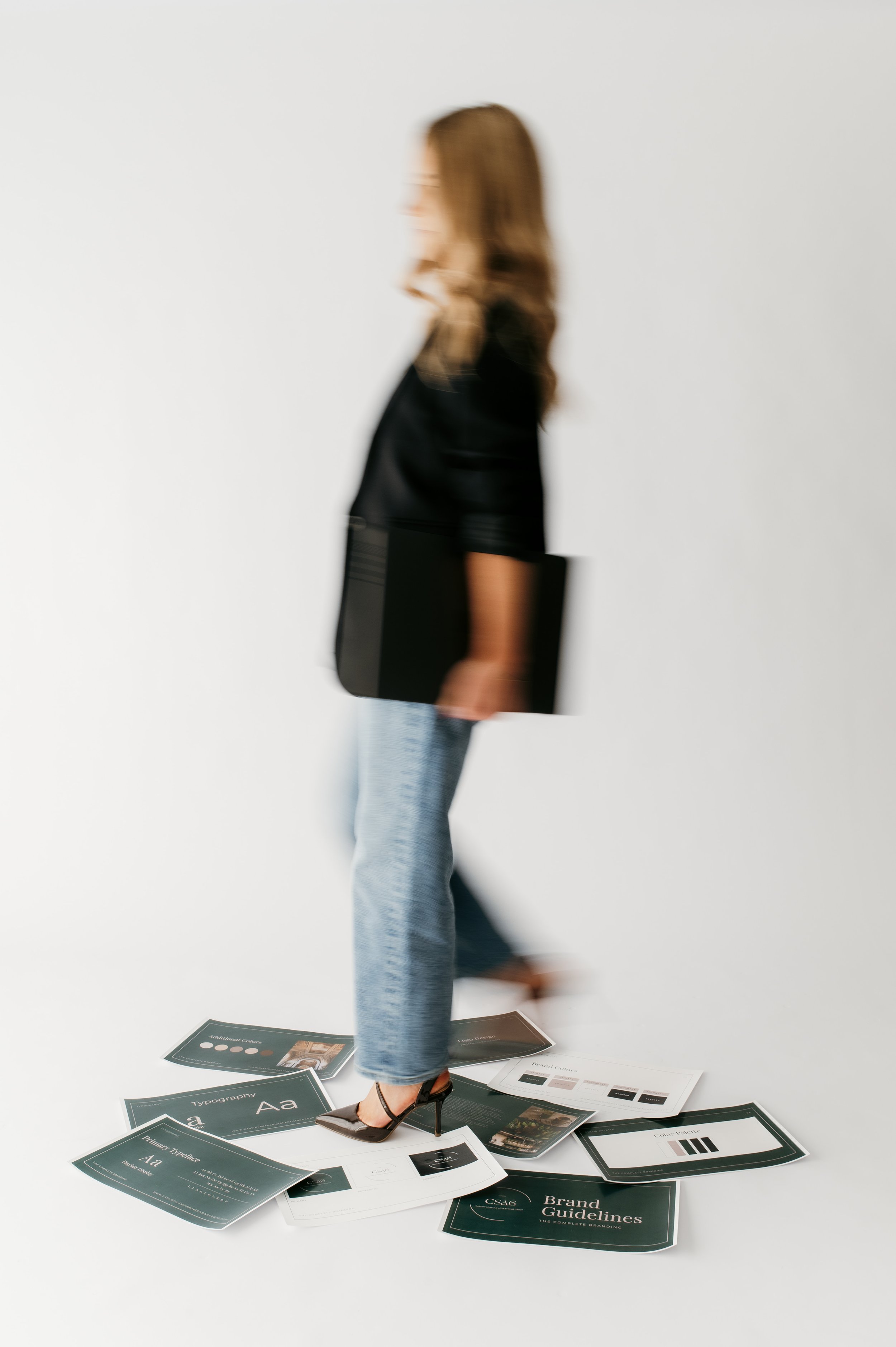 A woman in high heels and jeans walking over scattered presentation papers on a white floor, with a white background.