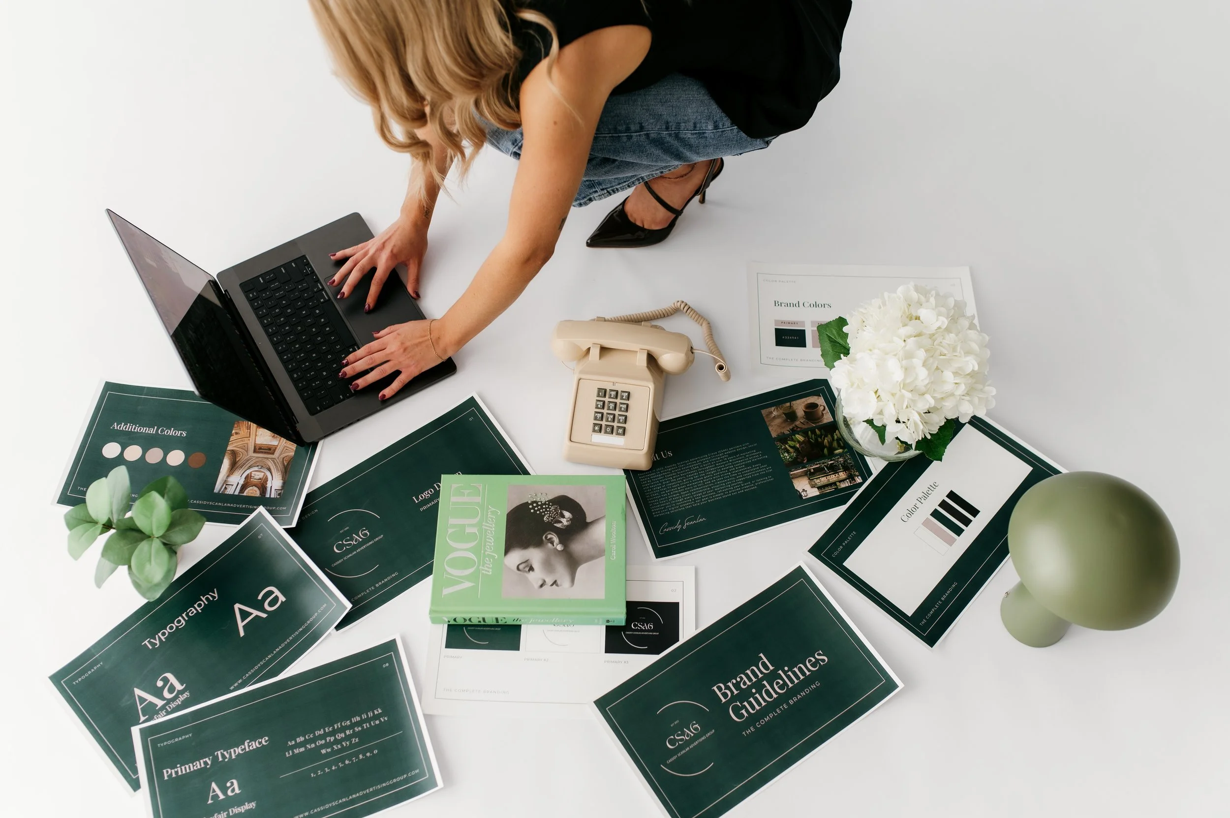 Person crouching on the floor working on a laptop surrounded by various design and branding materials, including color swatches, booklets, a vintage beige rotary phone, and a plant with white flowers.