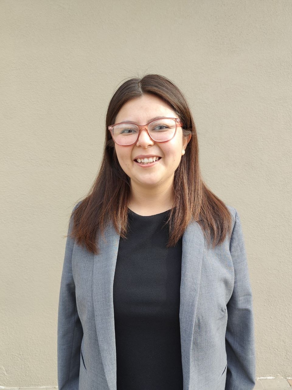 Mujer sonriendo con gafas, cabello castaño, vestida con blazer gris y camiseta negra, de pie frente a una pared beige.