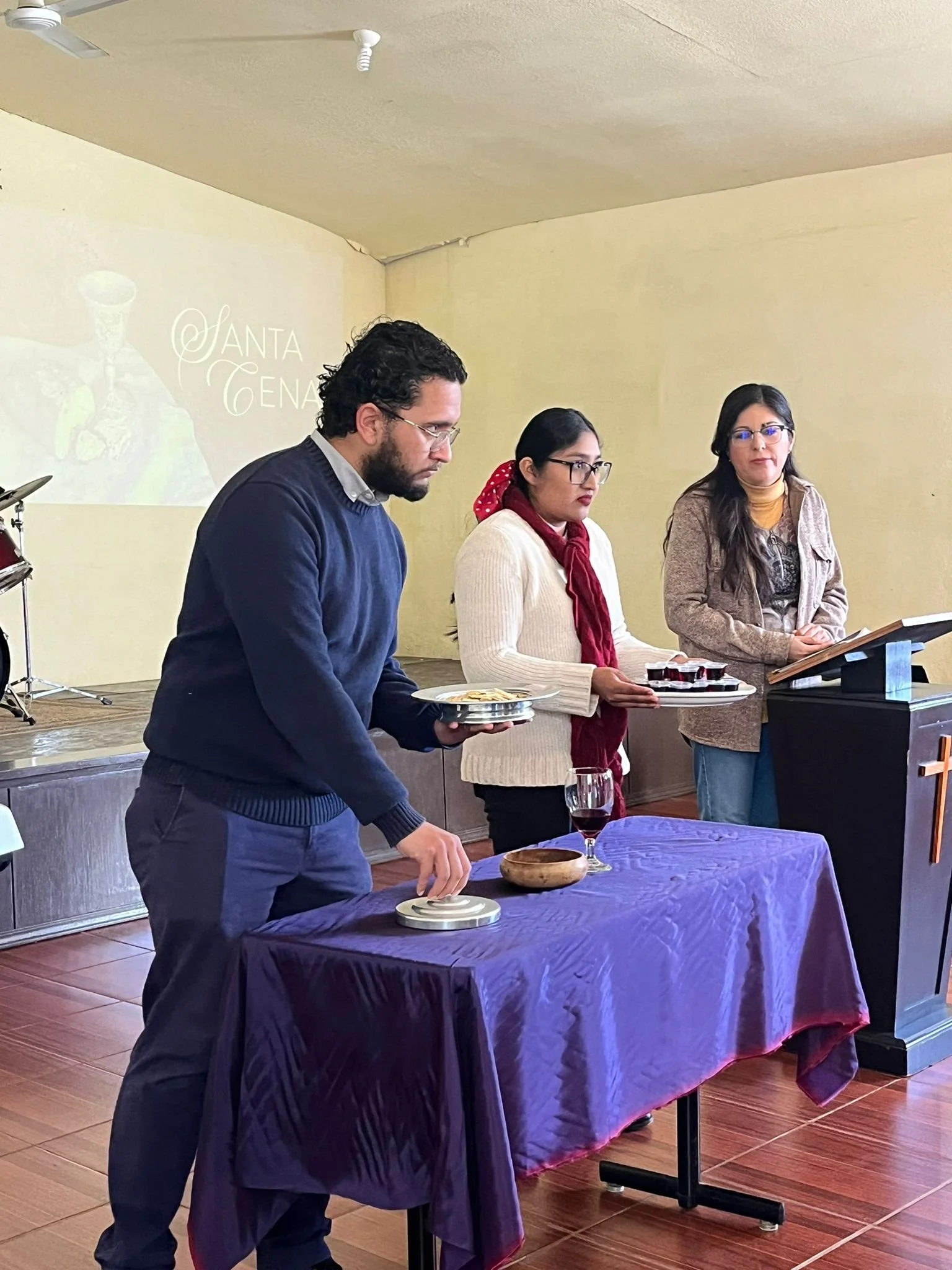 Tres personas participando en una ceremonia, con una de ellas tocando un vaso sobre una mesa cubierta con tela morada, en un espacio interior con paredes de color beige y piso de madera, en un evento formal o religioso.
