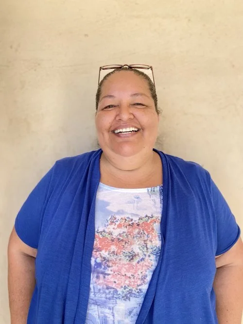 Mujer sonriendo con gafas sobre la cabeza, lleva una camiseta con un diseño y un suéter azul, fondo beige.