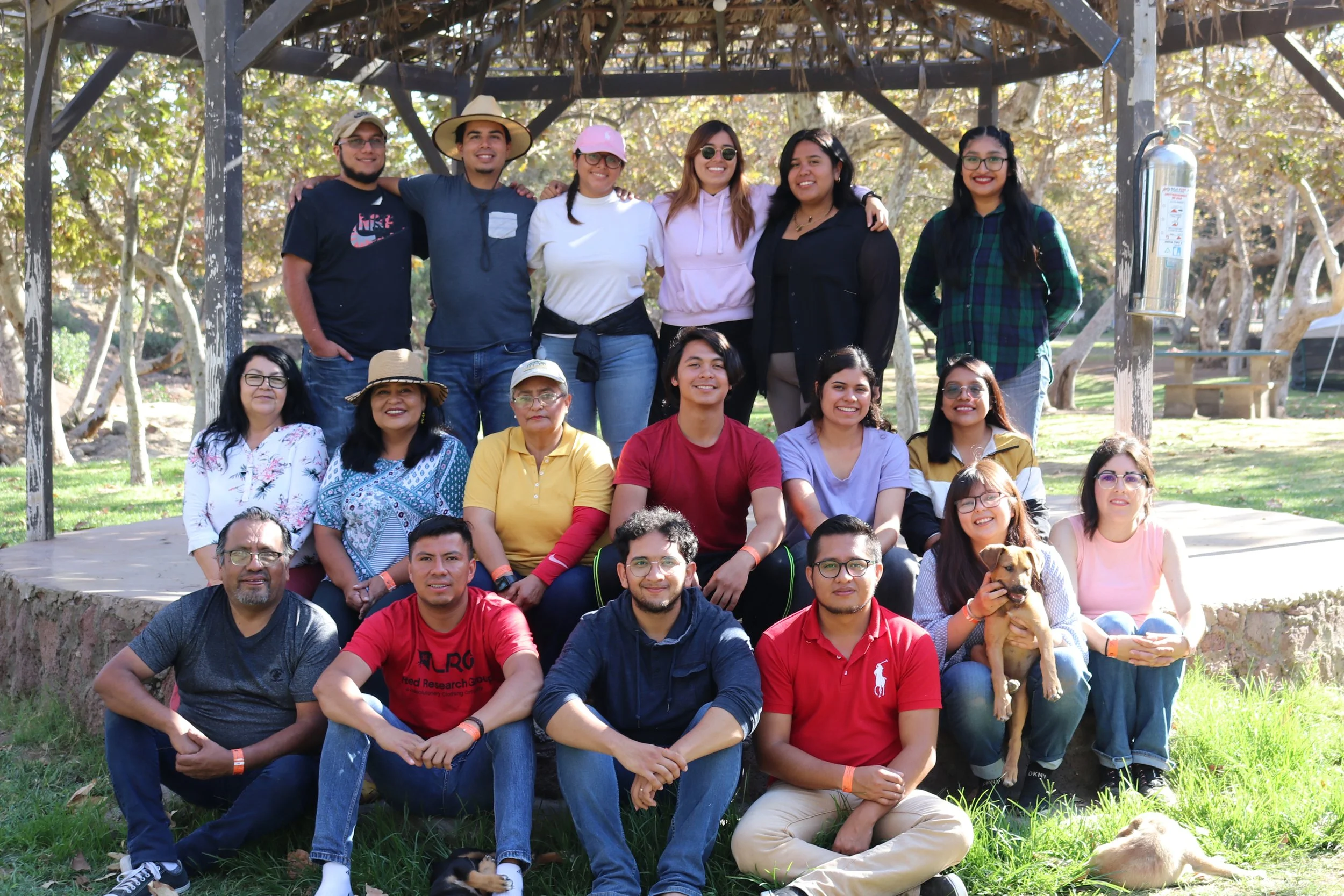Grupo de personas y perros en parque, algunos sonriendo, otros sentados o de pie, con árboles y estructura de madera en fondo.