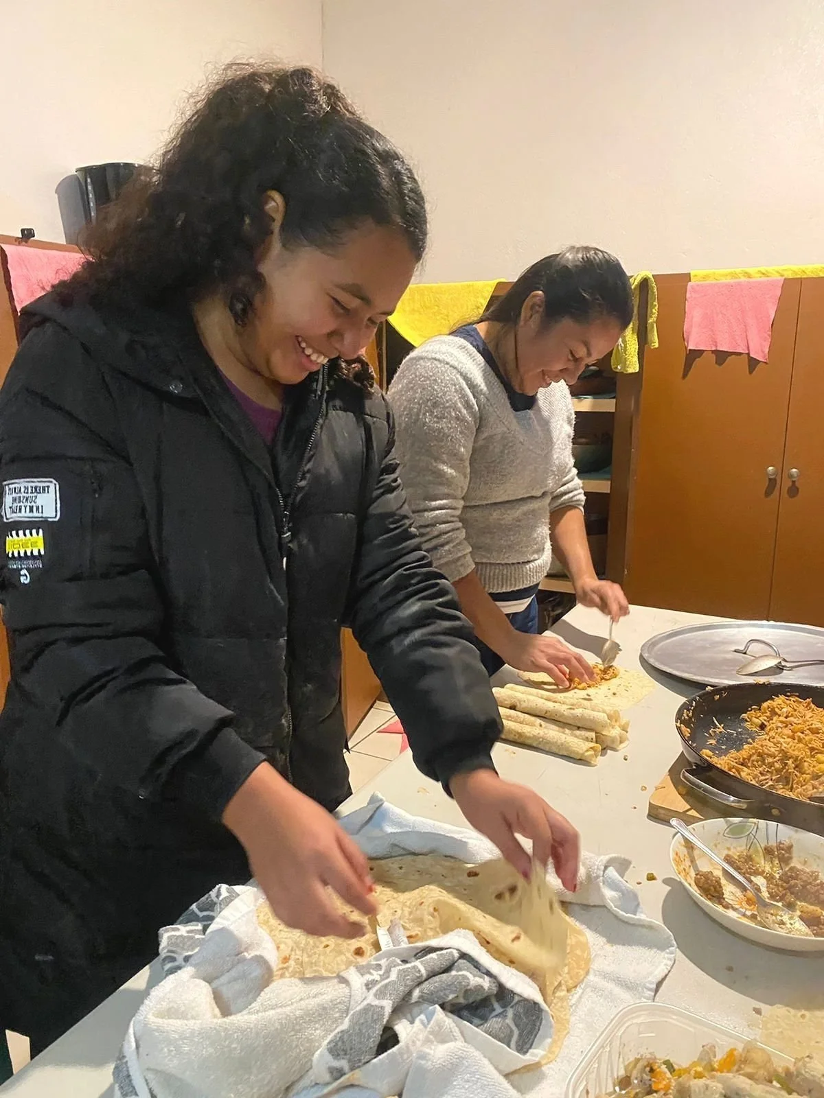 Dos mujeres preparando comida mexicana, colocando tortillas y rellenándolas con ingredientes en una mesa, en un ambiente casero.