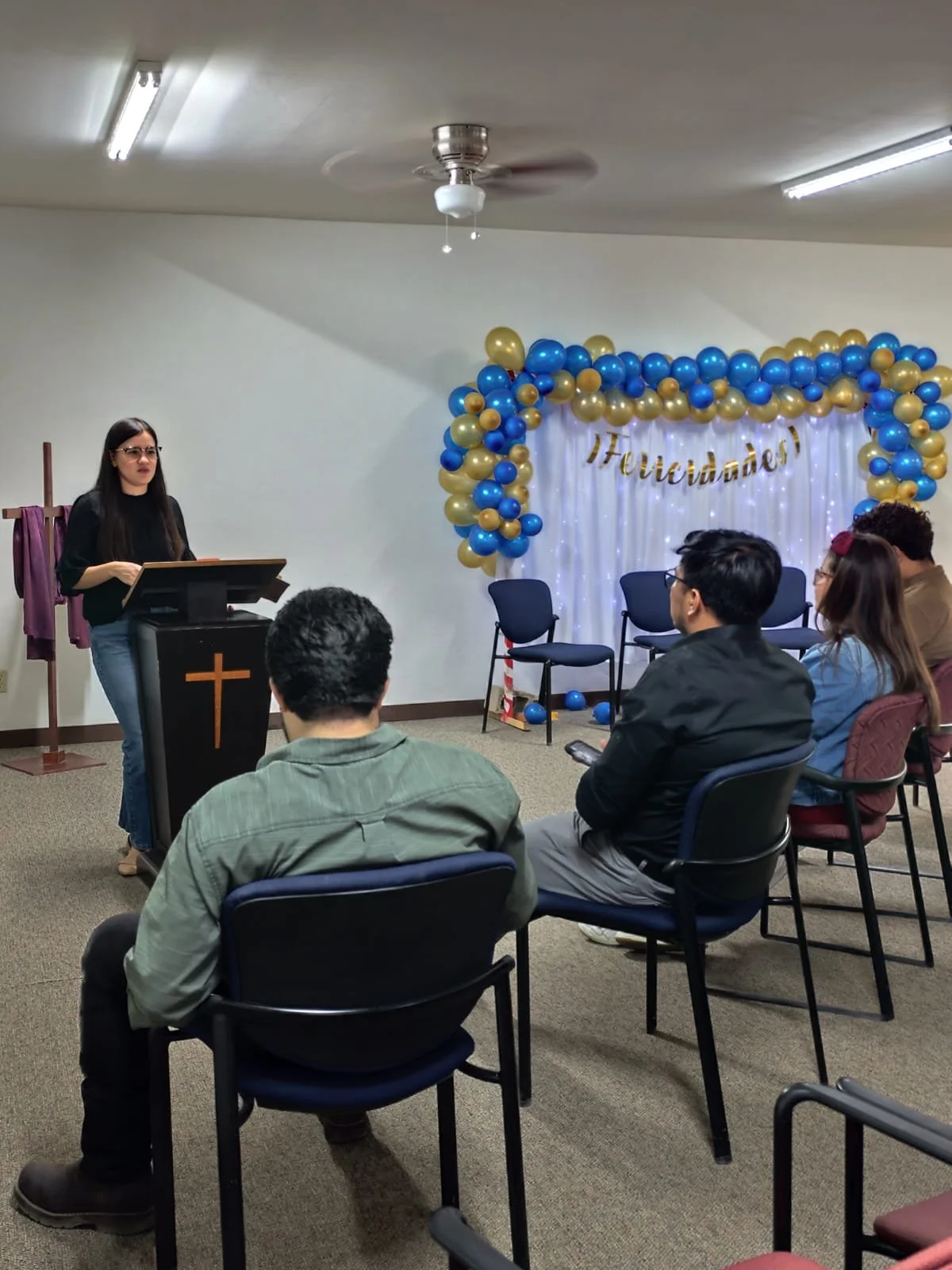 Una mujer habla en un podio con una cruz, mientras un grupo de personas escucha en una sala con decoración de globos azules y dorados y un cartel que dice '¡Felicidades!'.