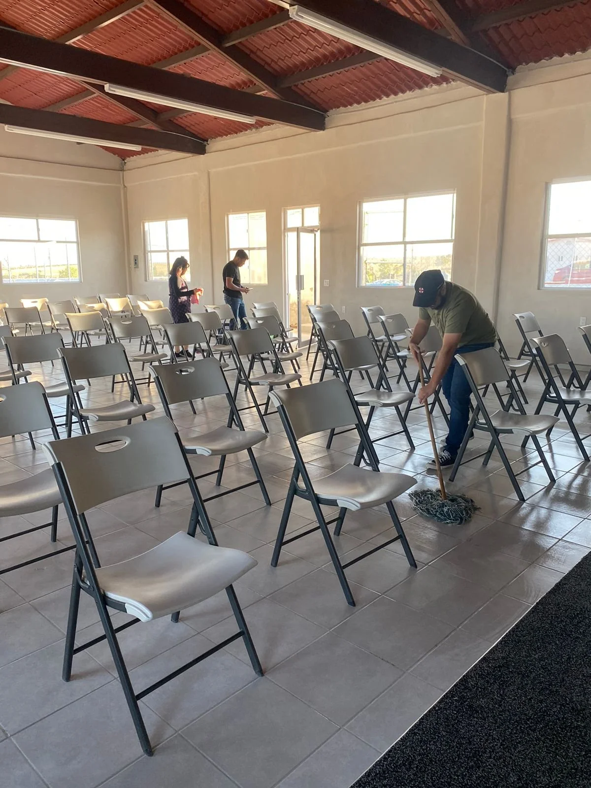 Varias personas limpiando sillas en un salón amplio con ventanas grandes y techo de madera. La sala está casi vacía y soleada.