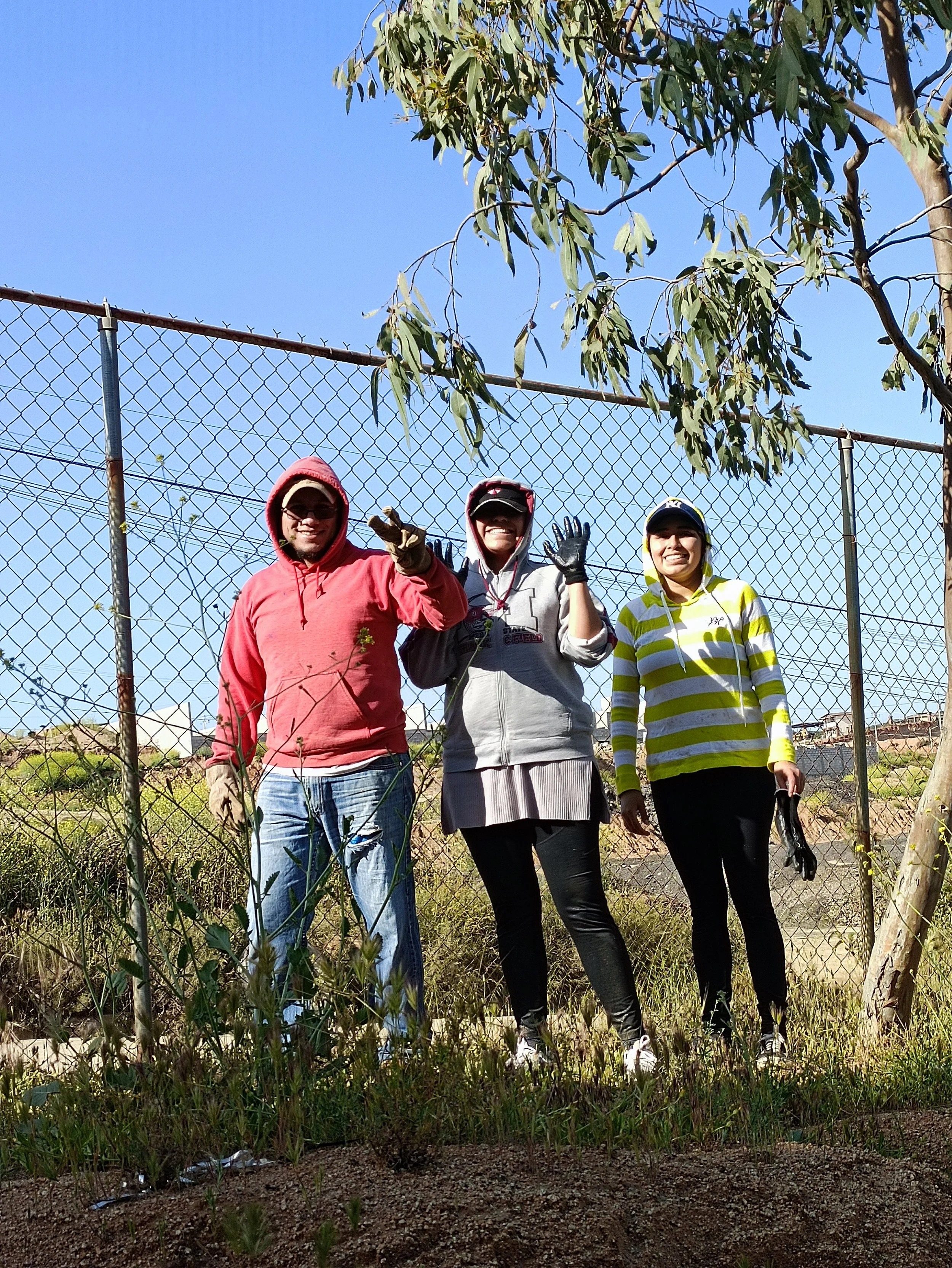 Tres personas sonrientes y vestidas con ropa de colores brillantes, de pie frente a una cerca de malla metálica, en un área con vegetación y árboles, en un día soleado con cielo despejado.