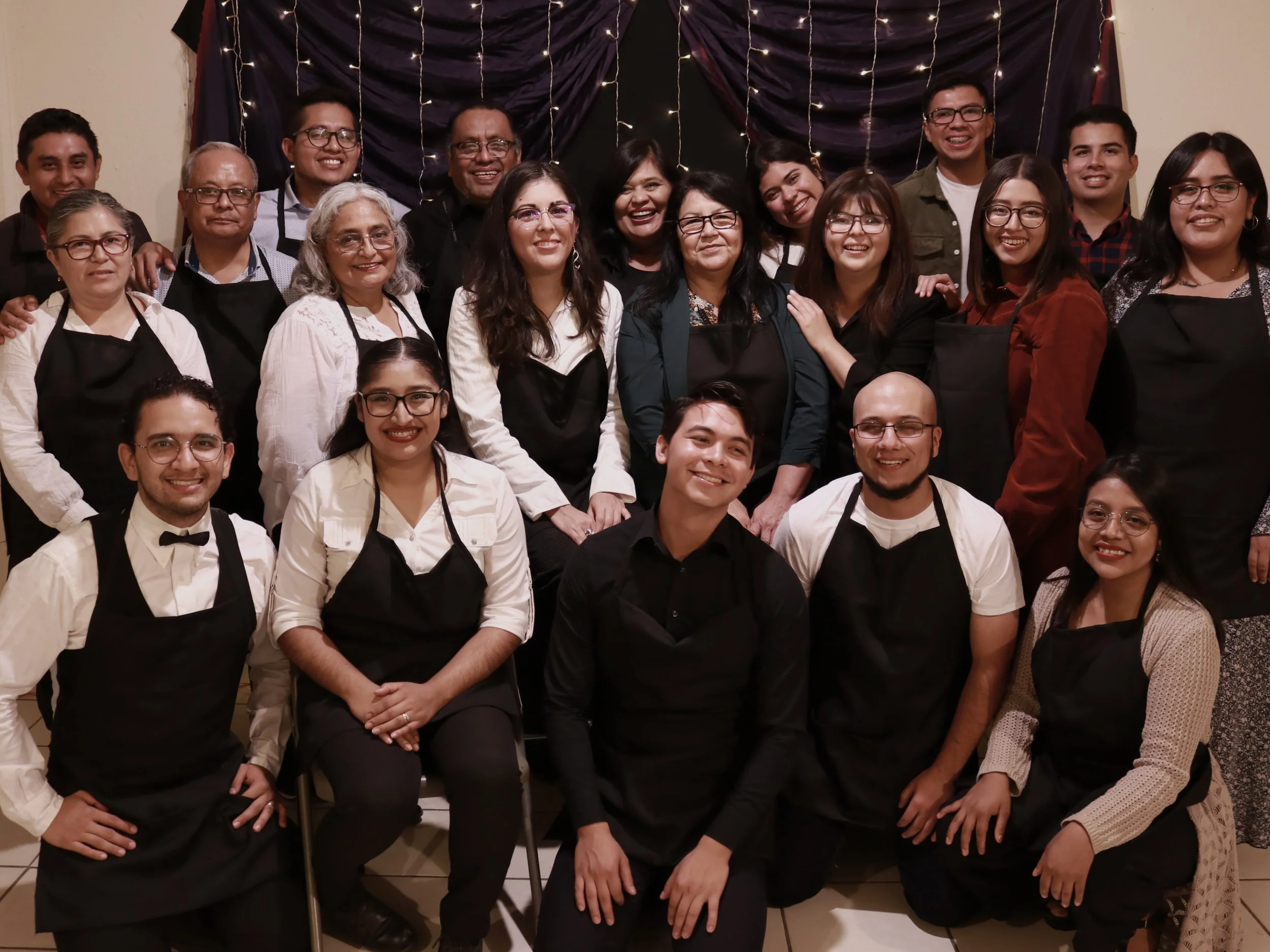 Grupo de personas sonriendo, algunas con delantales negros y otras con ropa casual, en una reunión o evento social, con cortinas oscuras y luces decorativas en el fondo.