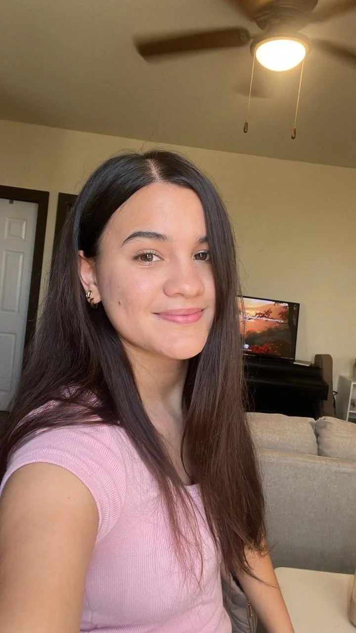 Joven mujer con cabello largo y oscuro, usando una camiseta rosa, sonriendo en un ambiente hogareño con televisión y sofá.