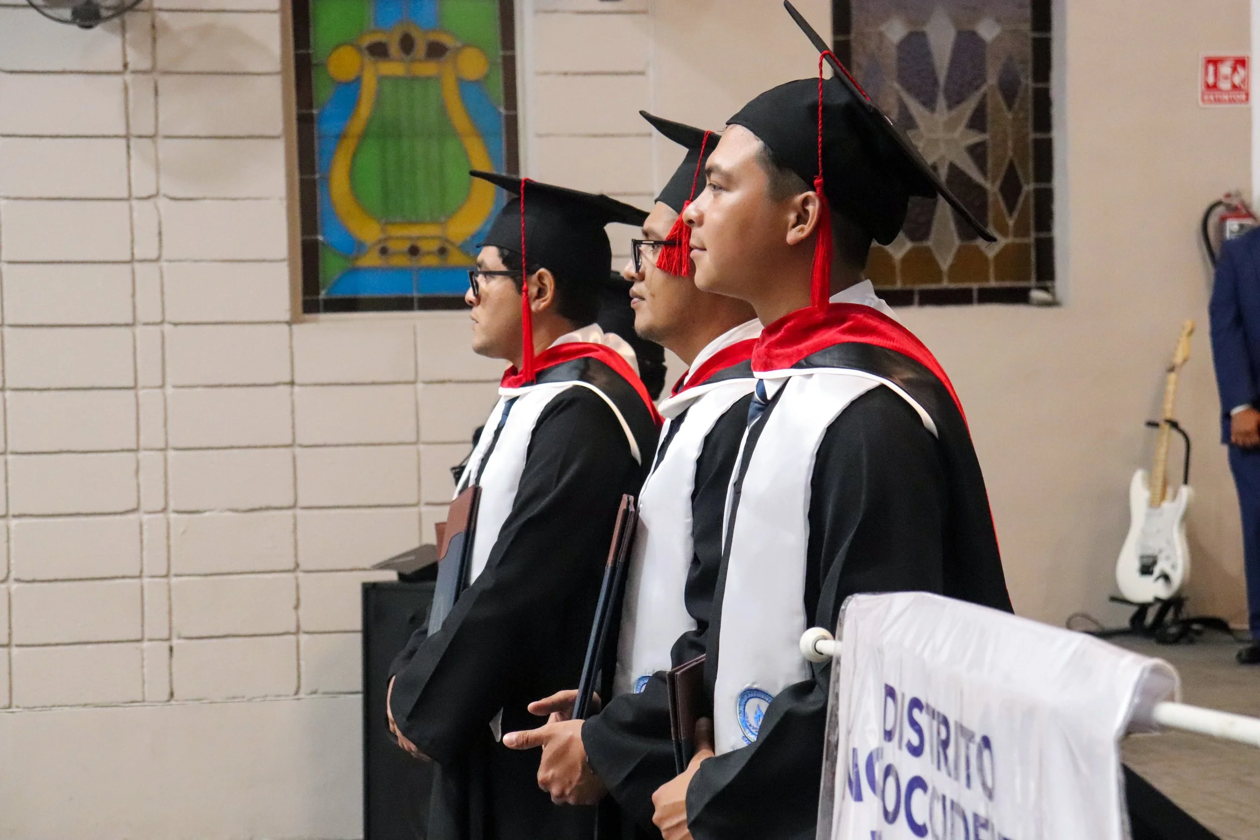 Cuatro graduados con toga y birrete en una ceremonia académica, de perfil, sentados en fila en un salón con paredes de ladrillo y ventanas decoradas con mosaicos de colores.