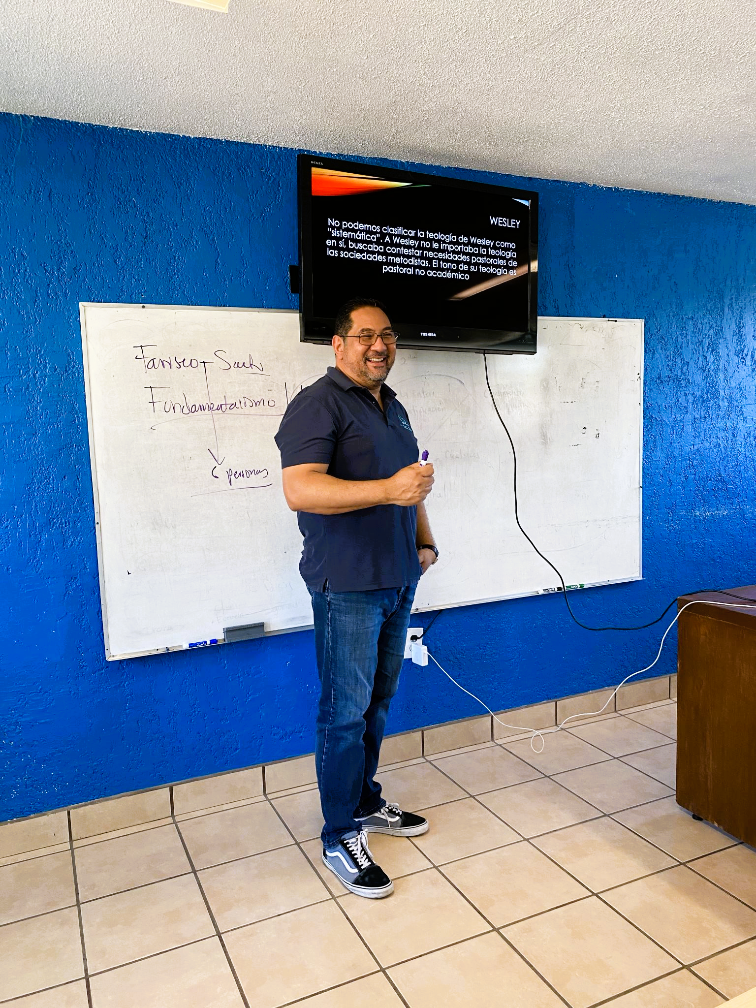 Hombre sonriente con gafas y barba, sosteniendo un marcador, frente a una pizarra blanca y una pantalla de televisión en una aula con paredes azules. La pizarra tiene escritos apuntes sobre fundamentos y personas, y la pantalla muestra texto en español.