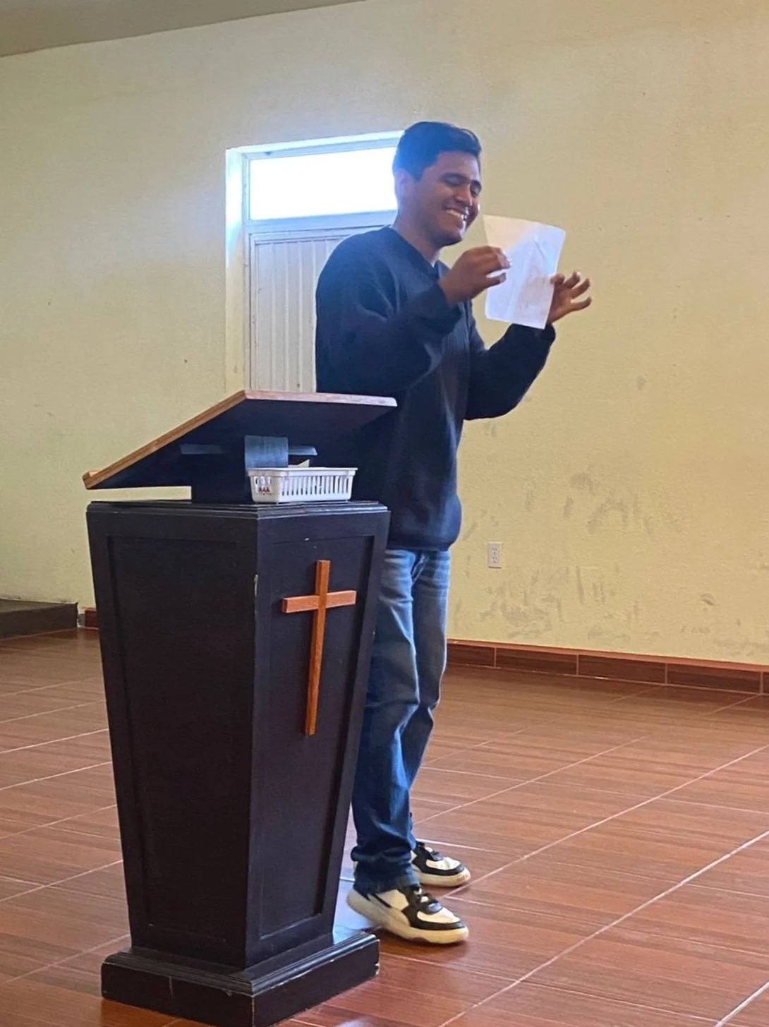 Un hombre sonriente de pie detrás de un púlpito con una cruz en una iglesia, sosteniendo un papel en la mano y parece estar en medio de un discurso o lectura.