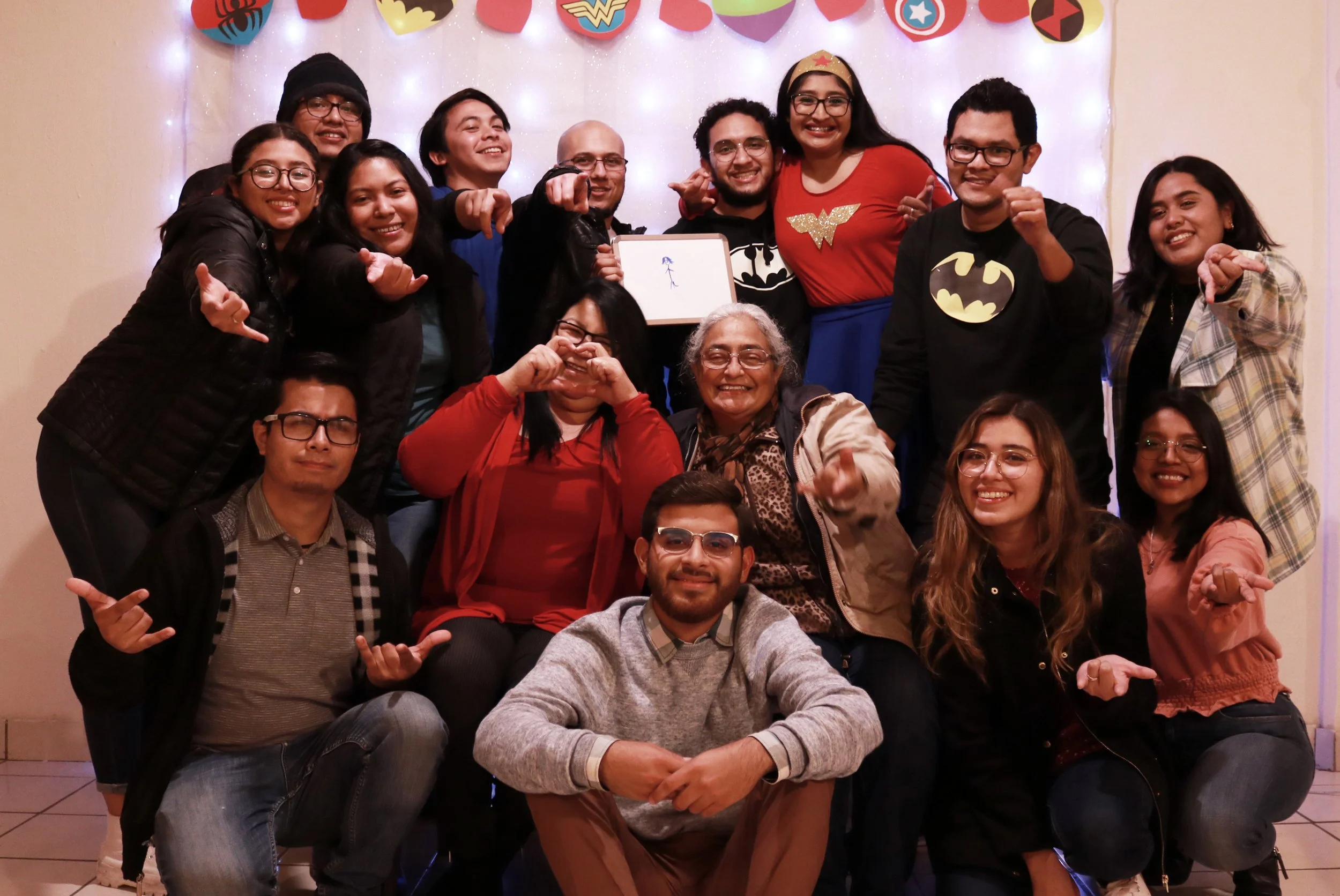 Un grupo de personas sonriendo y posando para una foto grupal en una fiesta con decoraciones de superhéroes en el fondo.