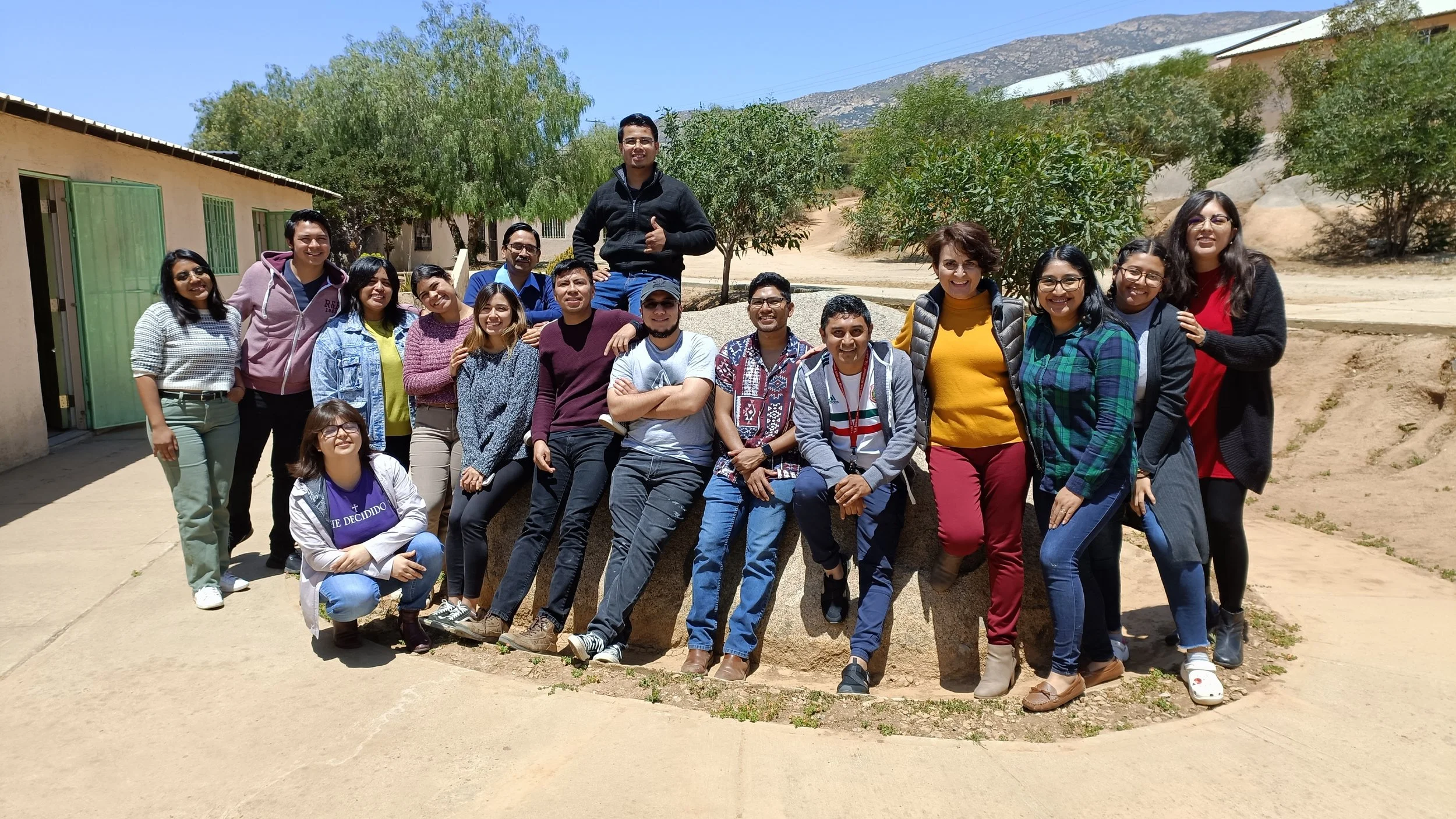 Un grupo de personas sonrientes posando al aire libre, con árboles y montañas de fondo, en un día soleado.