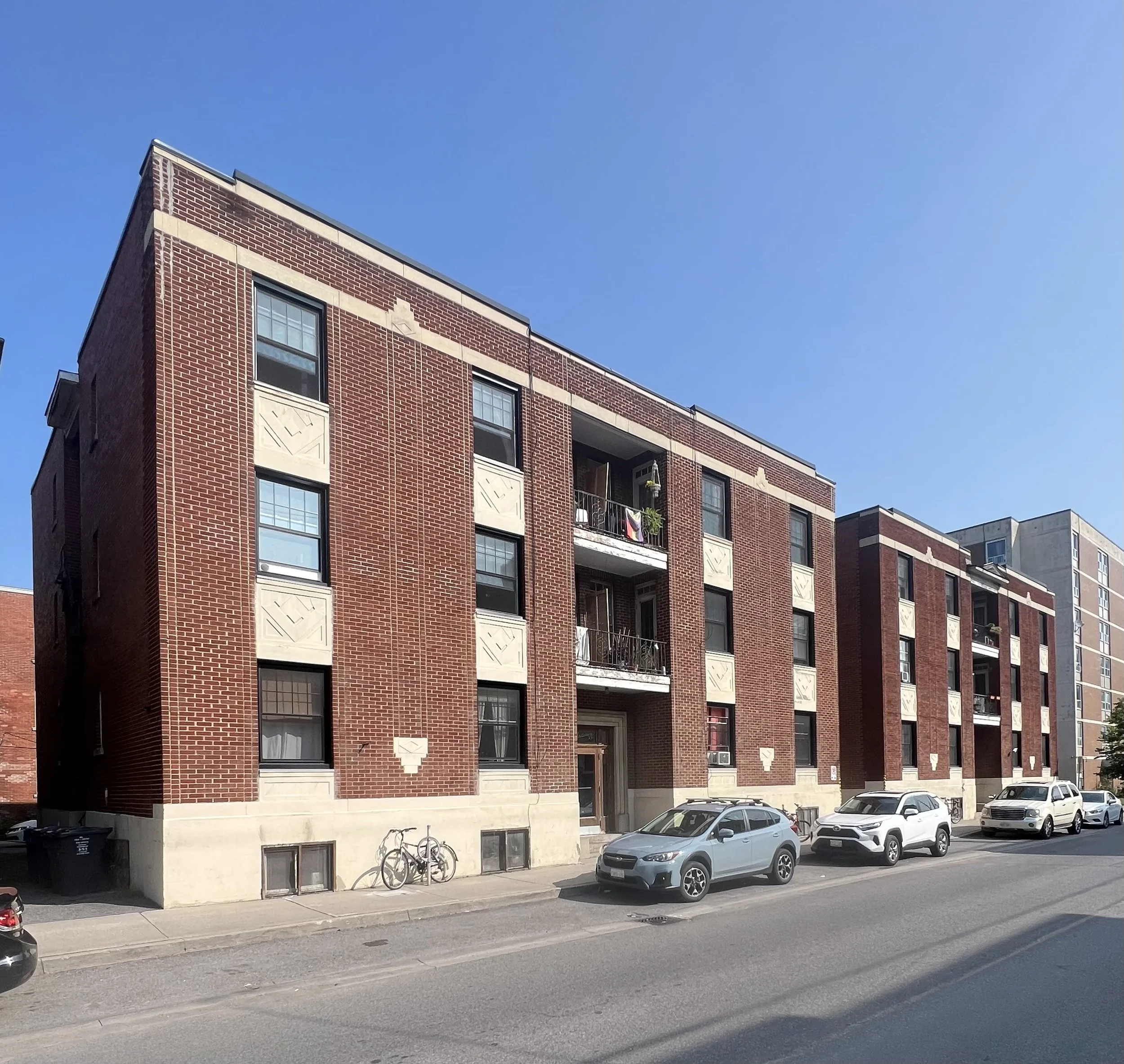 Three-story red brick apartment building with balconies on the second floor, parked cars in front, bicycles, and a clear blue sky.