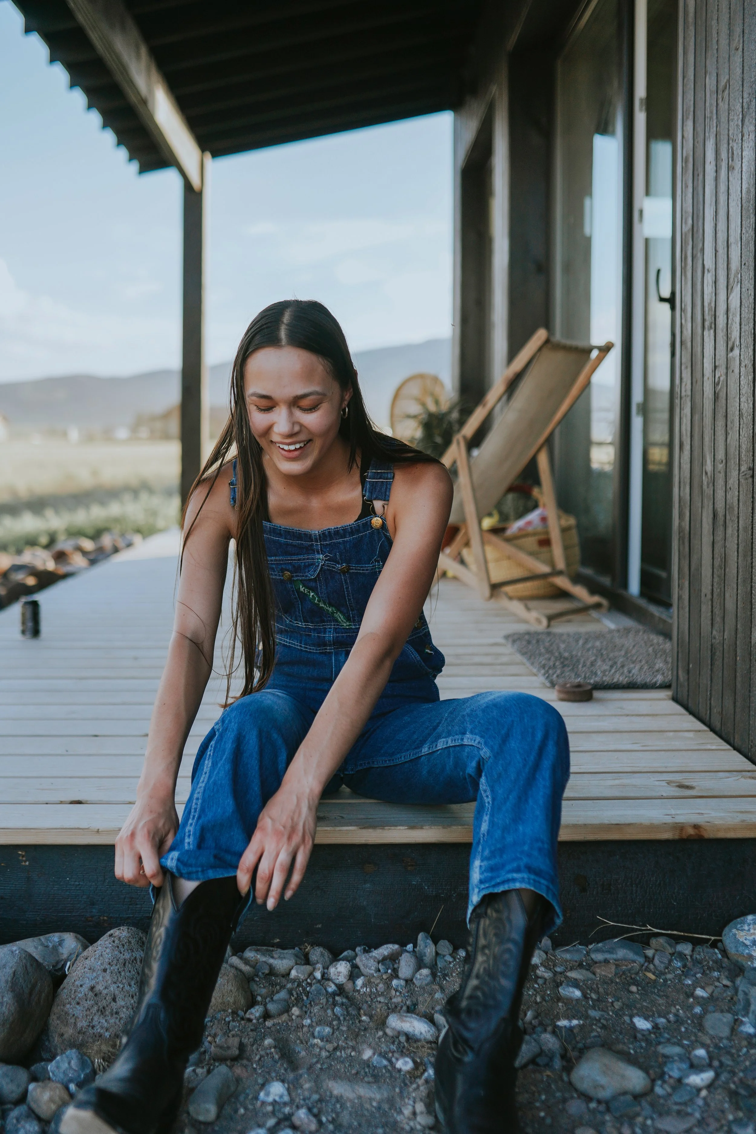 A photo of a smiling girl with long hair, sitting on a patio wearing vintage denim overalls and pulling on her cowboy boots with golden hour lighting.