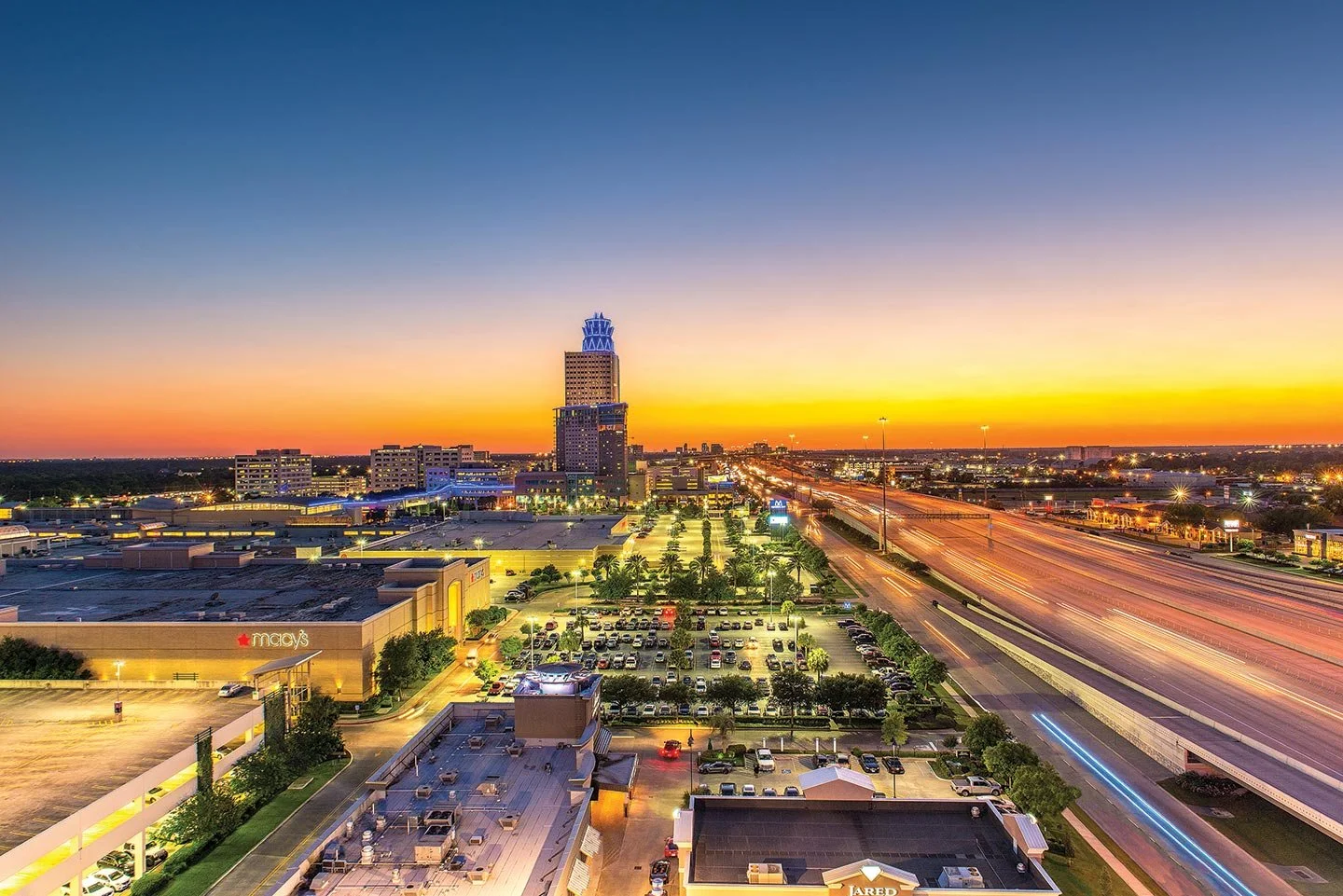 Cityscape at sunset in Houston, Texas, showing a skyline with a tall building and a mall parking lot, with streaks of car lights on nearby highways.
