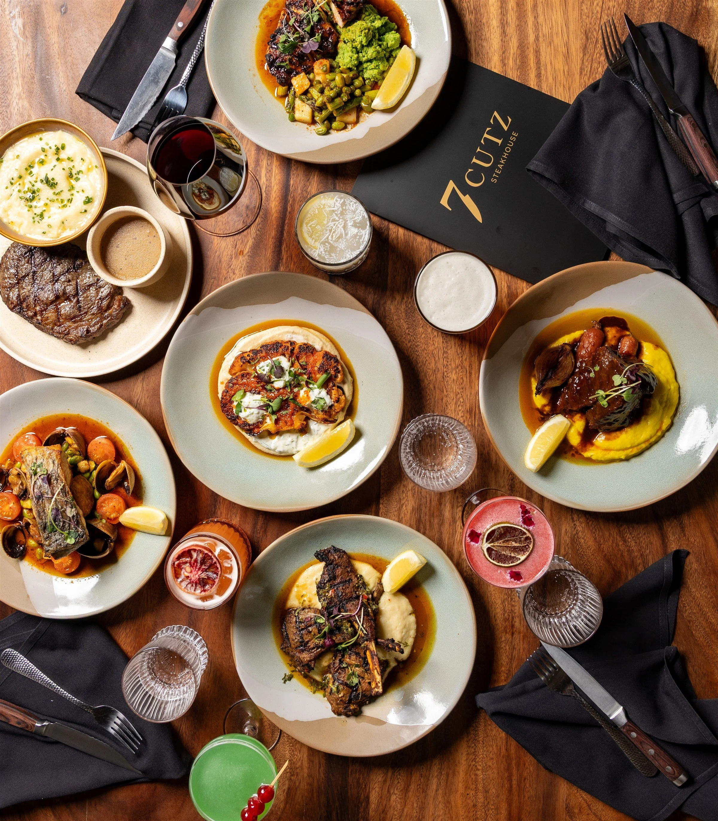 Overhead view of shared steak, lamb, fish and vegetarian dishes with cocktails on a timber table at 7 Cutz Steakhouse Canberra