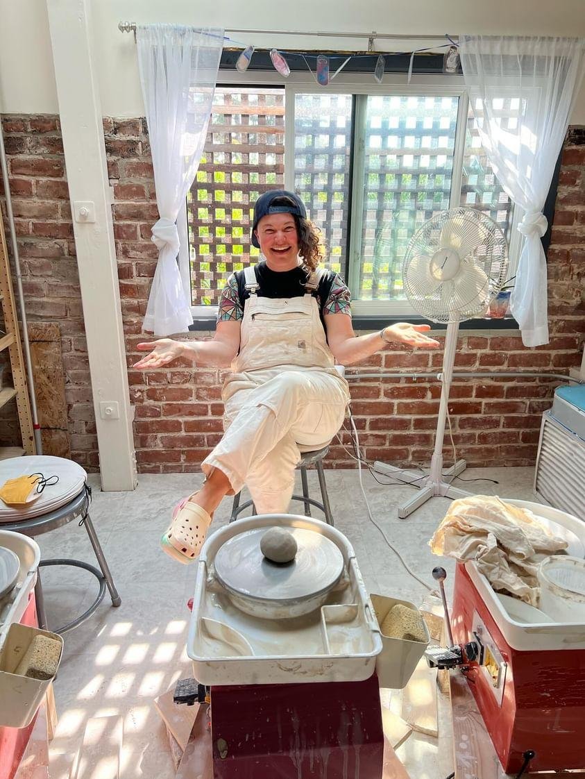 A kind, enthusiastic pottery teacher sits in front of a potter's wheel in a sunny clay studio