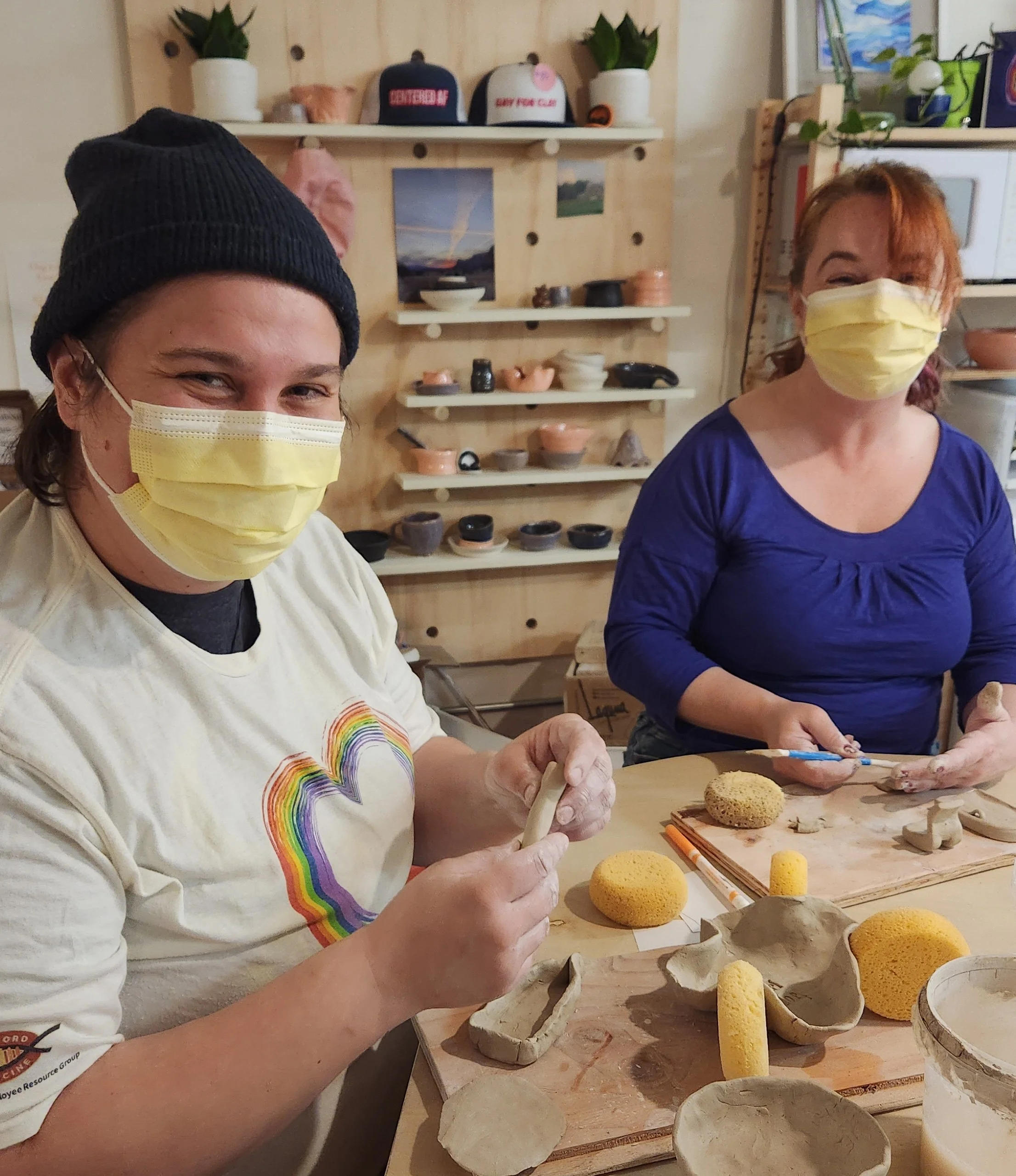 Two new friends are wearing masks and working on handbuilding clay projects at a welcoming pottery studio