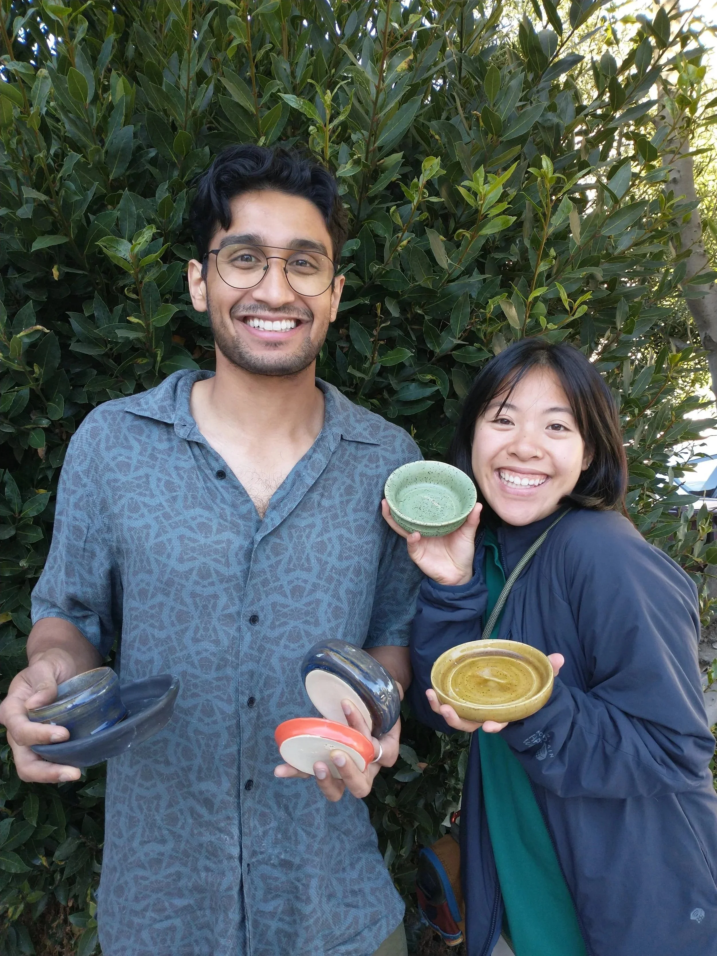 Two people holding ceramic bowls in front of a leafy background in the Bay Area. The left person holds several bowls, while the right person holds two, smiling at the camera.
