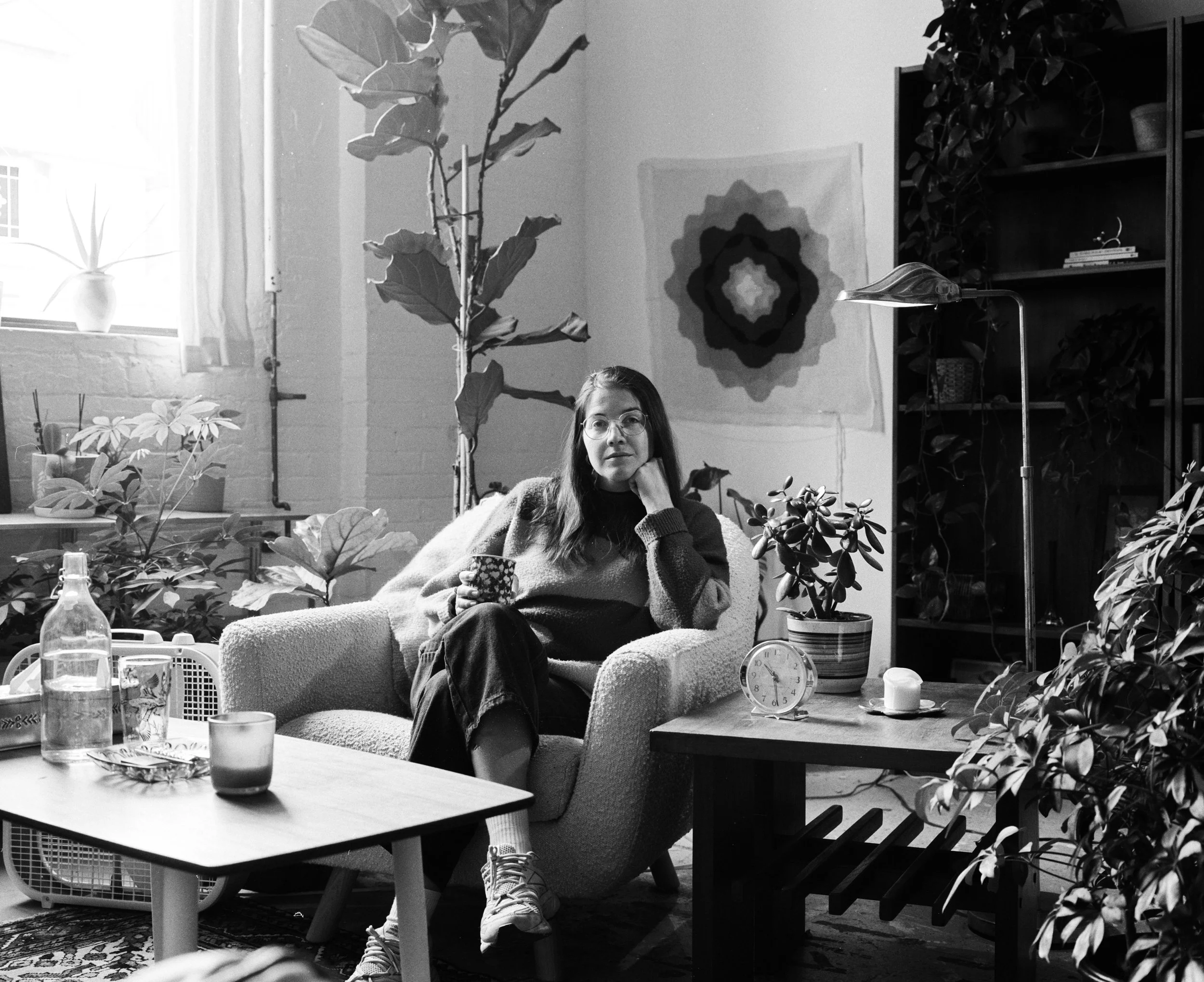 A woman with glasses sitting on a cozy armchair in a plant-filled room, holding a mug while looking at the camera. There are multiple potted plants, a clock, and a lamp on side tables, with a tapestry on the wall. Sunlight streams through a window.