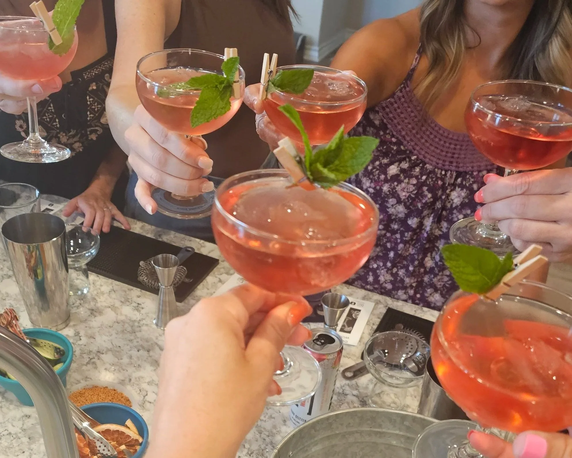 Multiple women are holding pink cocktails garnished with mint leaves in a lively social setting. The drinks are in coupe glasses, and the scene appears to be at a bar or a party with various bar tools and ingredients visible on the counter.