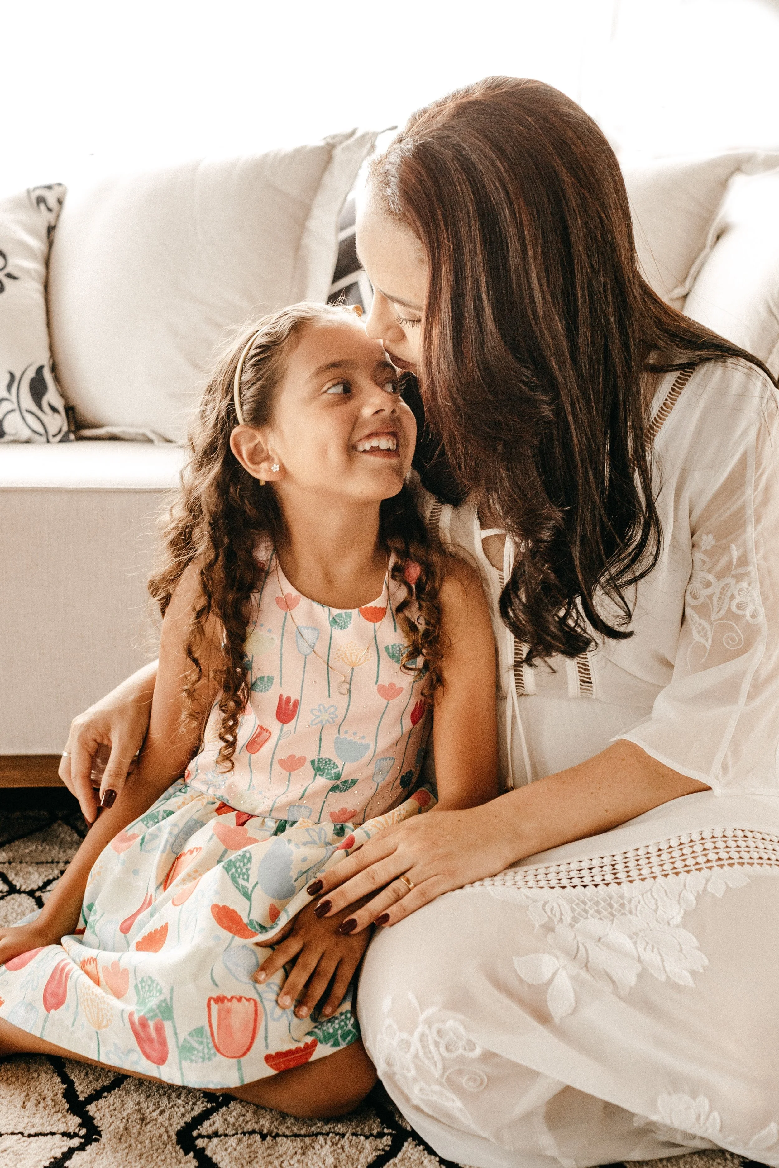 An adult woman and young girl sitting close together on the floor in a living room, sharing a joyful moment with their faces touching and smiling at each other.