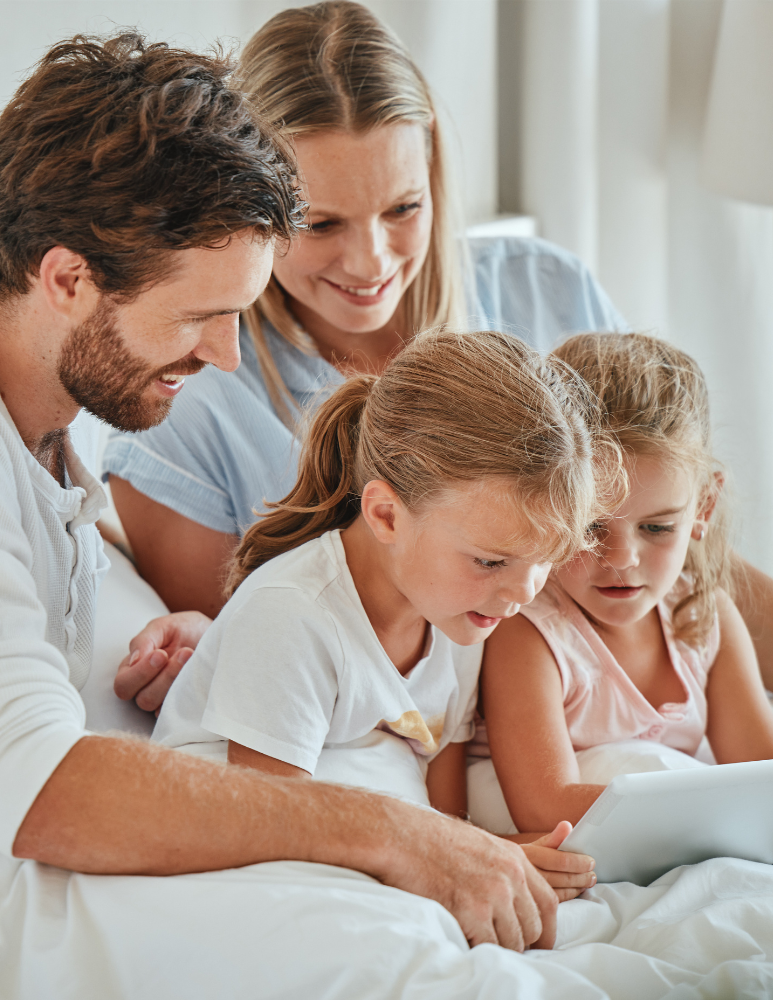 Family of four, two adults and two young girls, sitting close together on a bed looking at a tablet screen, smiling and engaged.