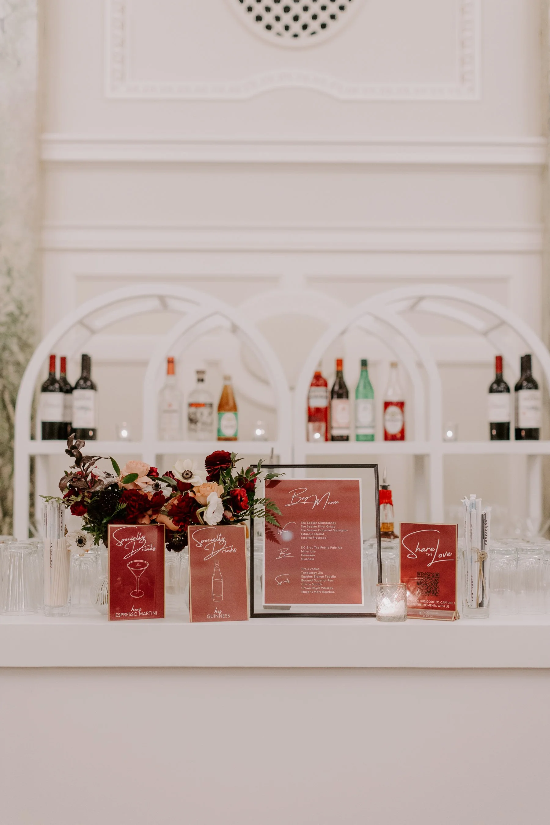 Bar signage in different shades of red and pink sit on the bar at a DC or Baltimore micro wedding cocktail hour. Drinks sit behind the bar and the bar is white.