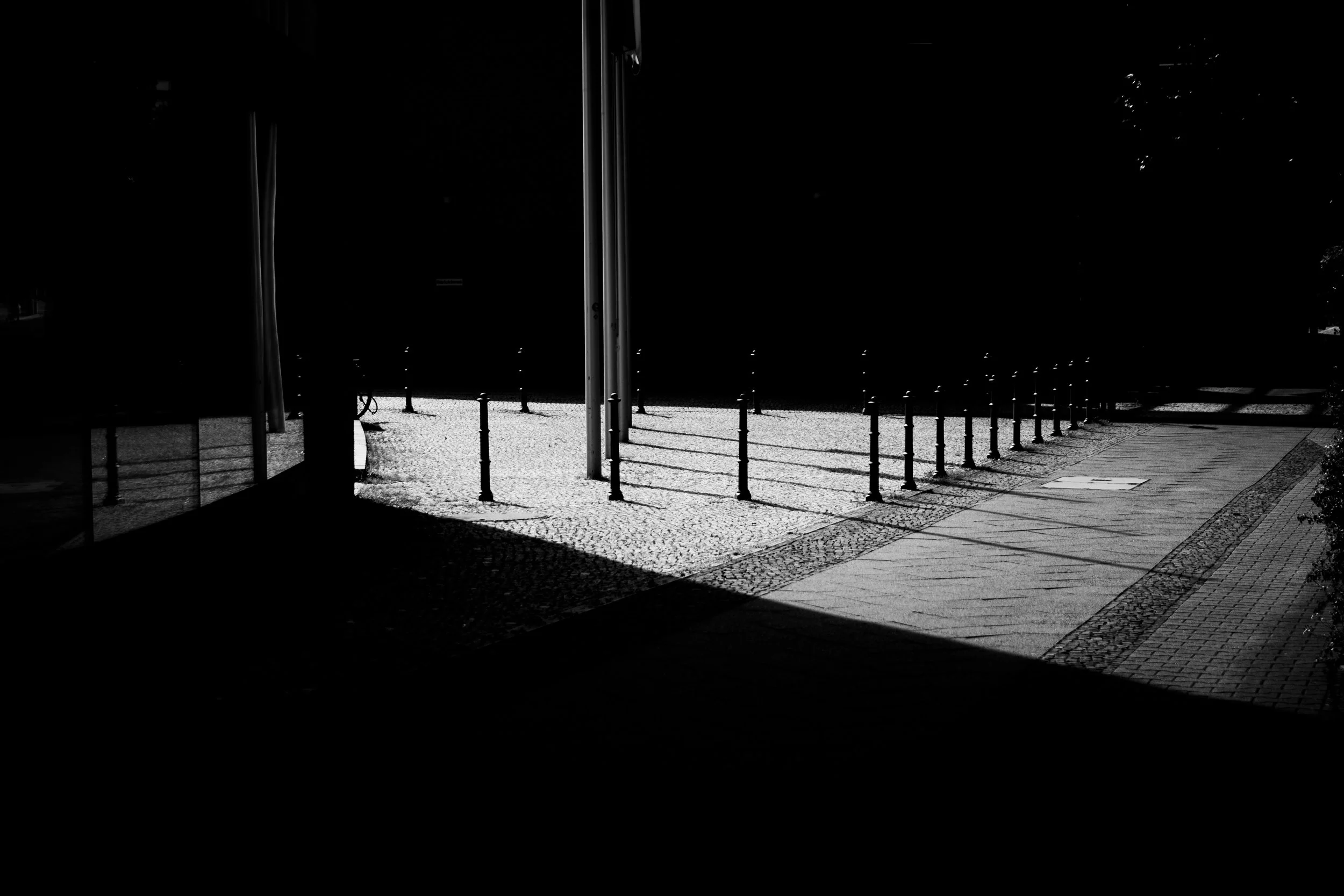 A black and white photograph of a sidewalk with a row of bollards, casting shadows, next to a glass building reflecting part of the scene.