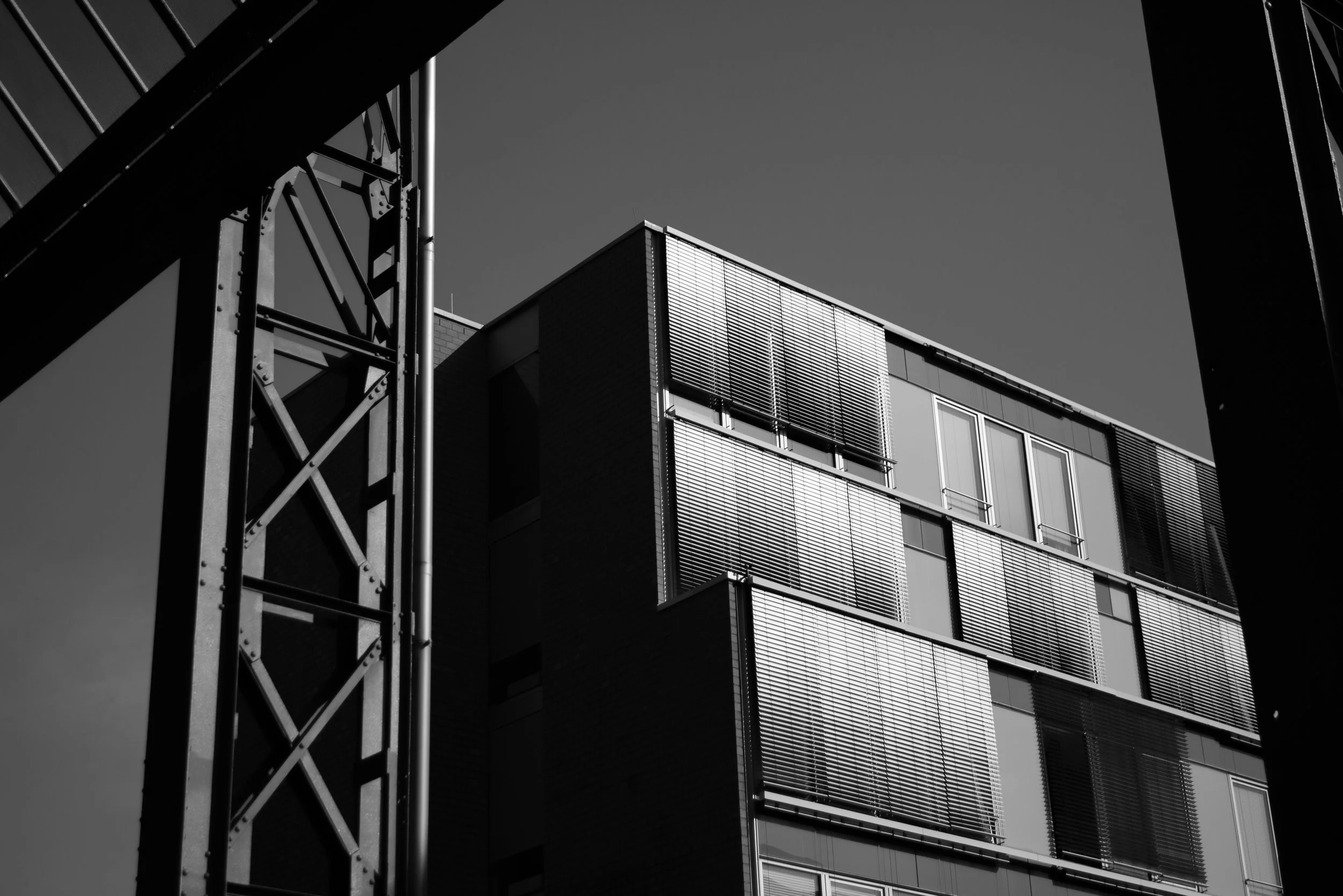 Black and white photo of modern building with large windows and metal blinds, framed by a metal structure in the foreground.