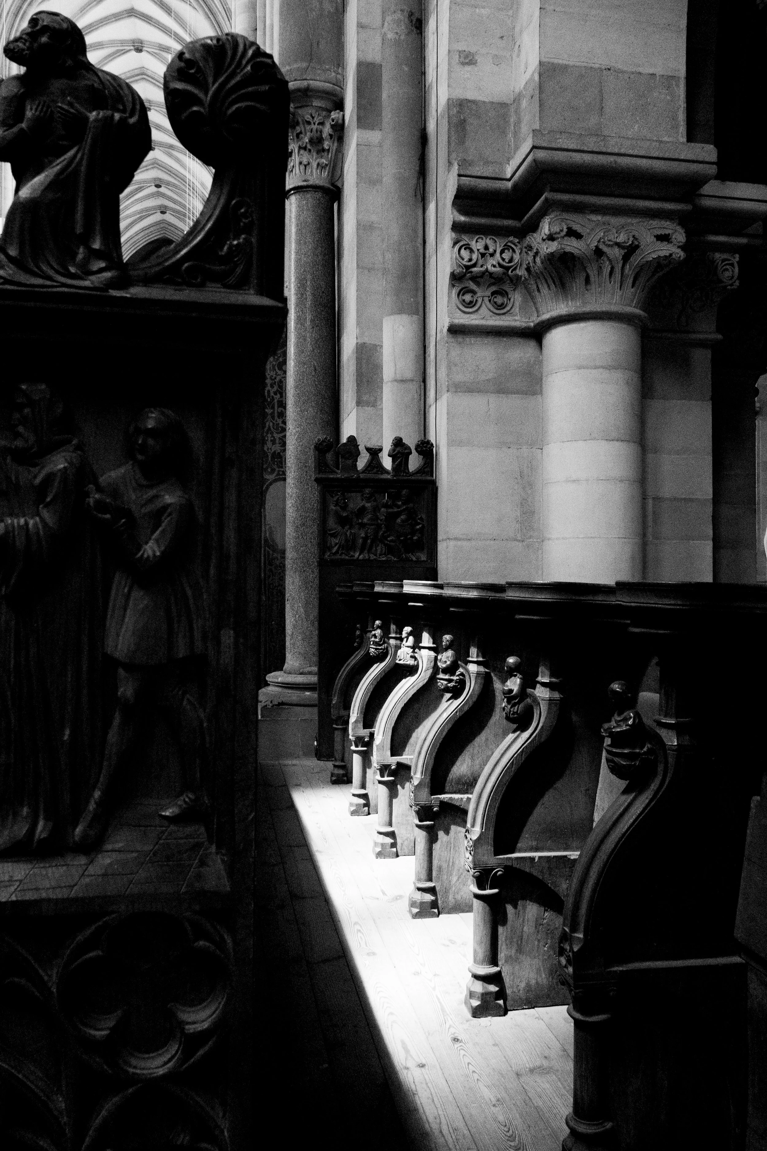 Black and white photo of ornate wooden choir stalls with carved figures, set against stone columns and walls inside a church.