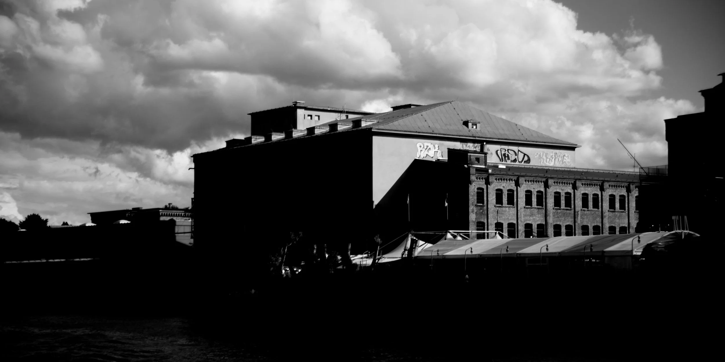 Black and white photo of buildings under a cloudy sky, with some graffiti on the upper part of one building.