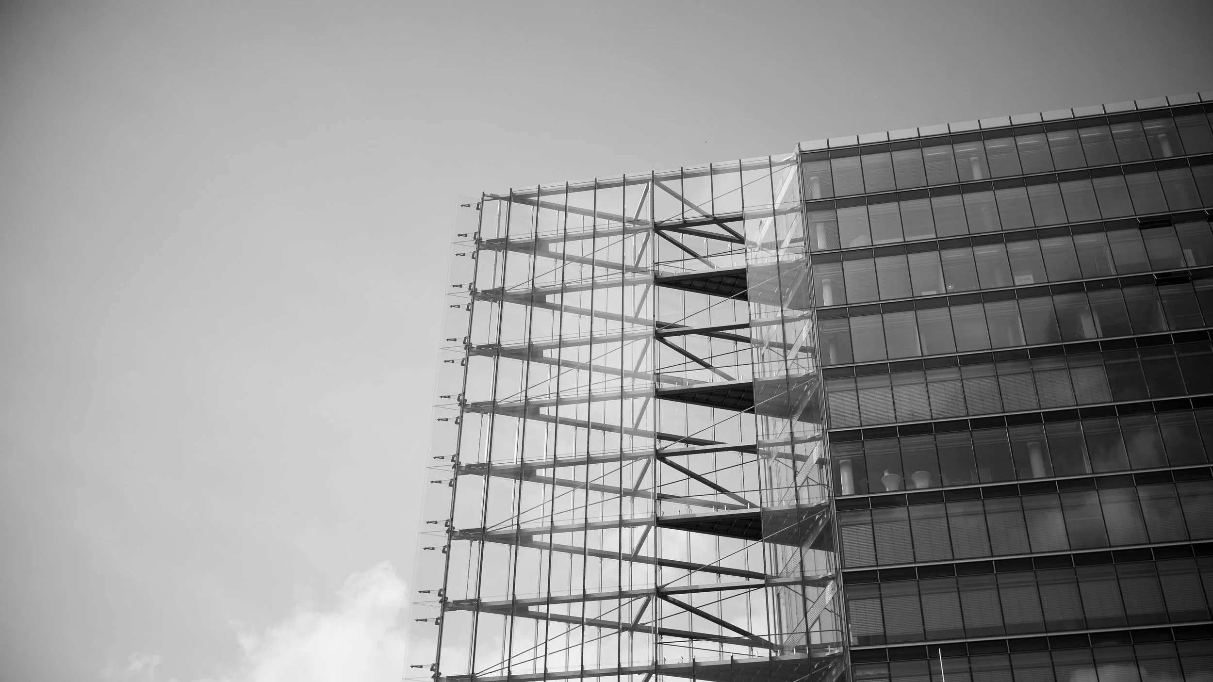 A black and white photo of a modern building with glass windows and a visible external skeleton or scaffolding structure on the left side.