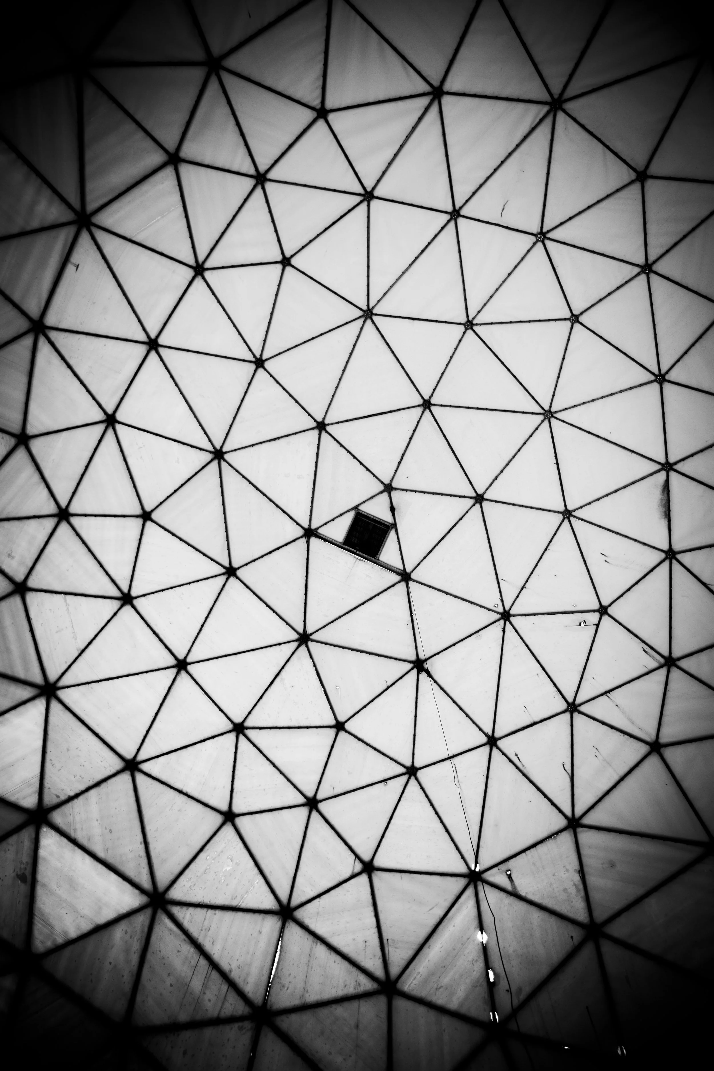 Interior view of a geodesic dome ceiling with a central opening, showing a grid of triangular panels and a small window.