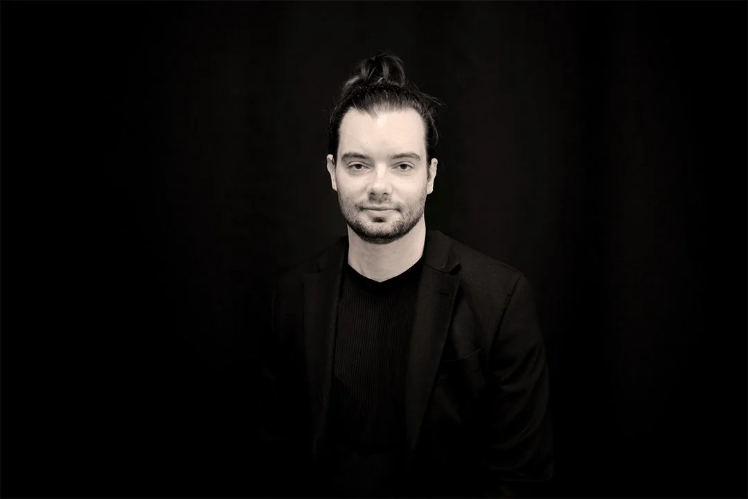 Black and white portrait of a man with long hair tied in a bun, wearing a black blazer and shirt, against a dark background.