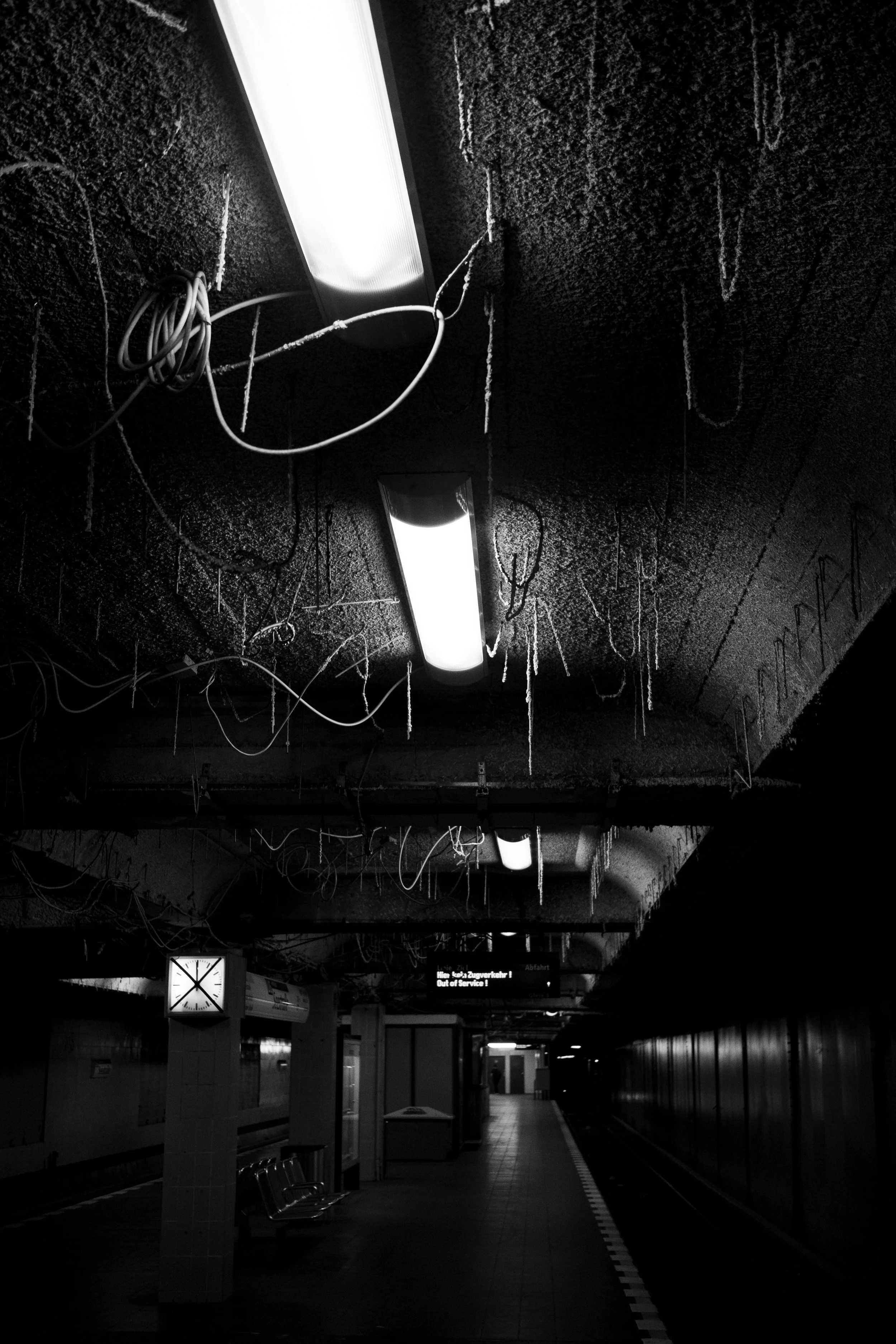A black and white photo of an underground subway station with exposed wiring hanging from the ceiling and fluorescent lights. The platform is empty with a row of seats and an electronic sign in the distance.