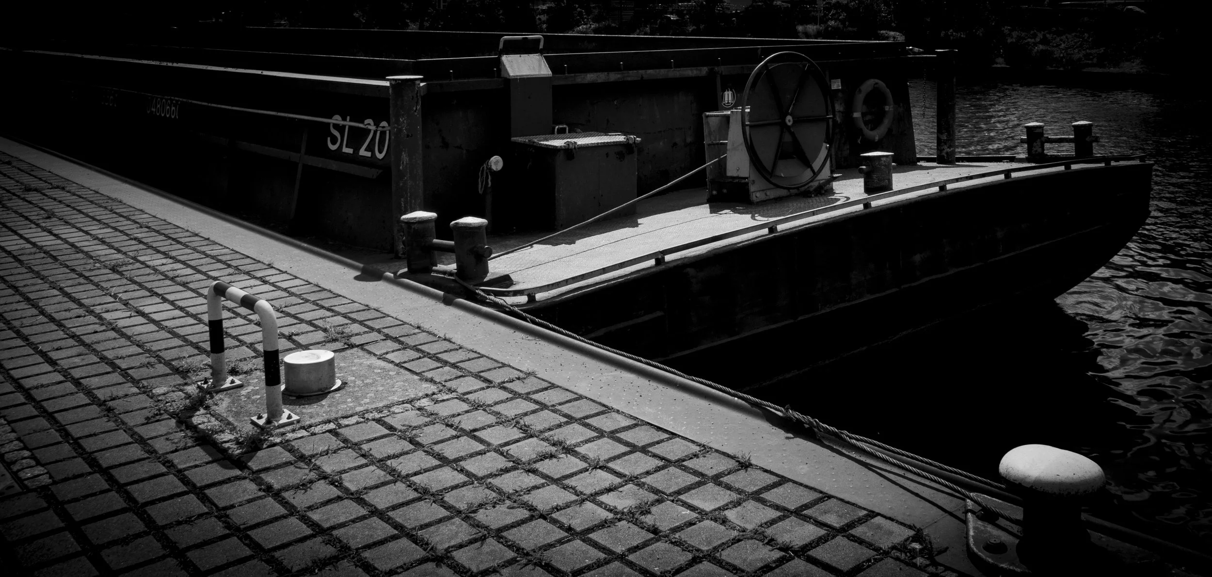 Black and white photo of a docked boat at a waterfront, with cobblestone pavement, bollards, and a mooring rope.