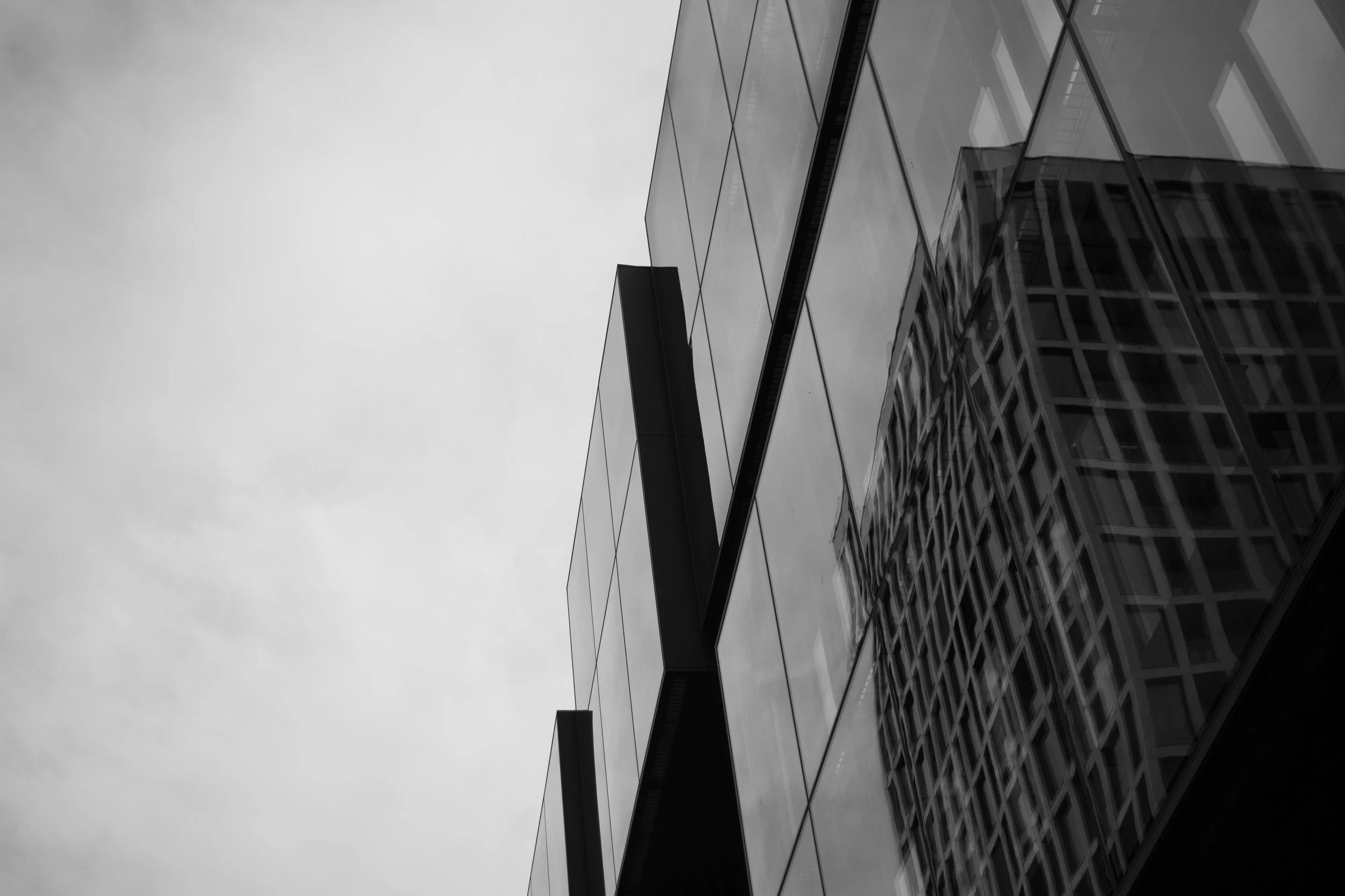 Black and white photo of modern glass building reflecting clouds and other buildings, with an overcast sky.