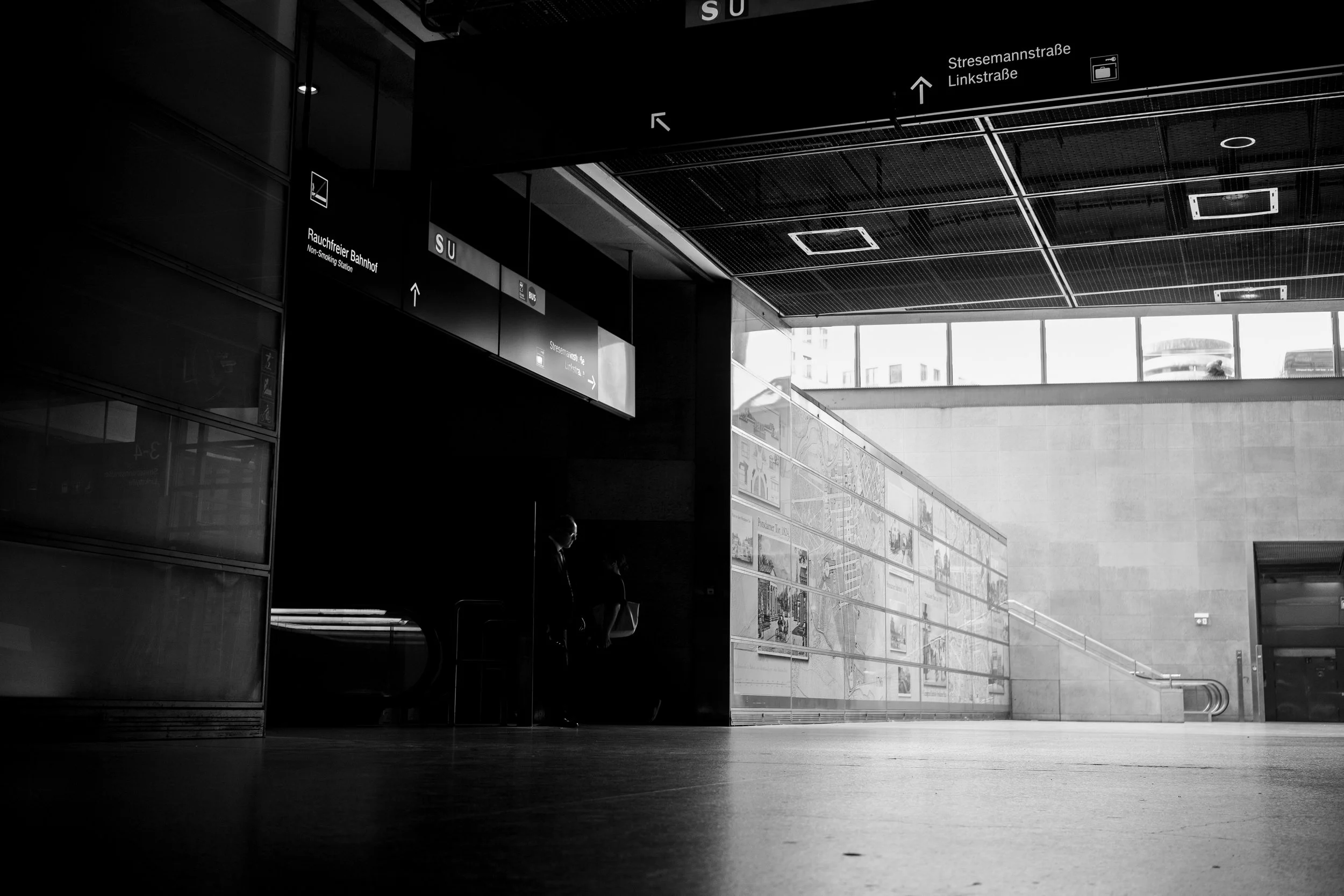 Black and white photo of an indoor train or subway station entrance with signage and a staircase leading upstairs. Two figures are sitting in the shadowed area, and sunlight is coming through large windows in the background.
