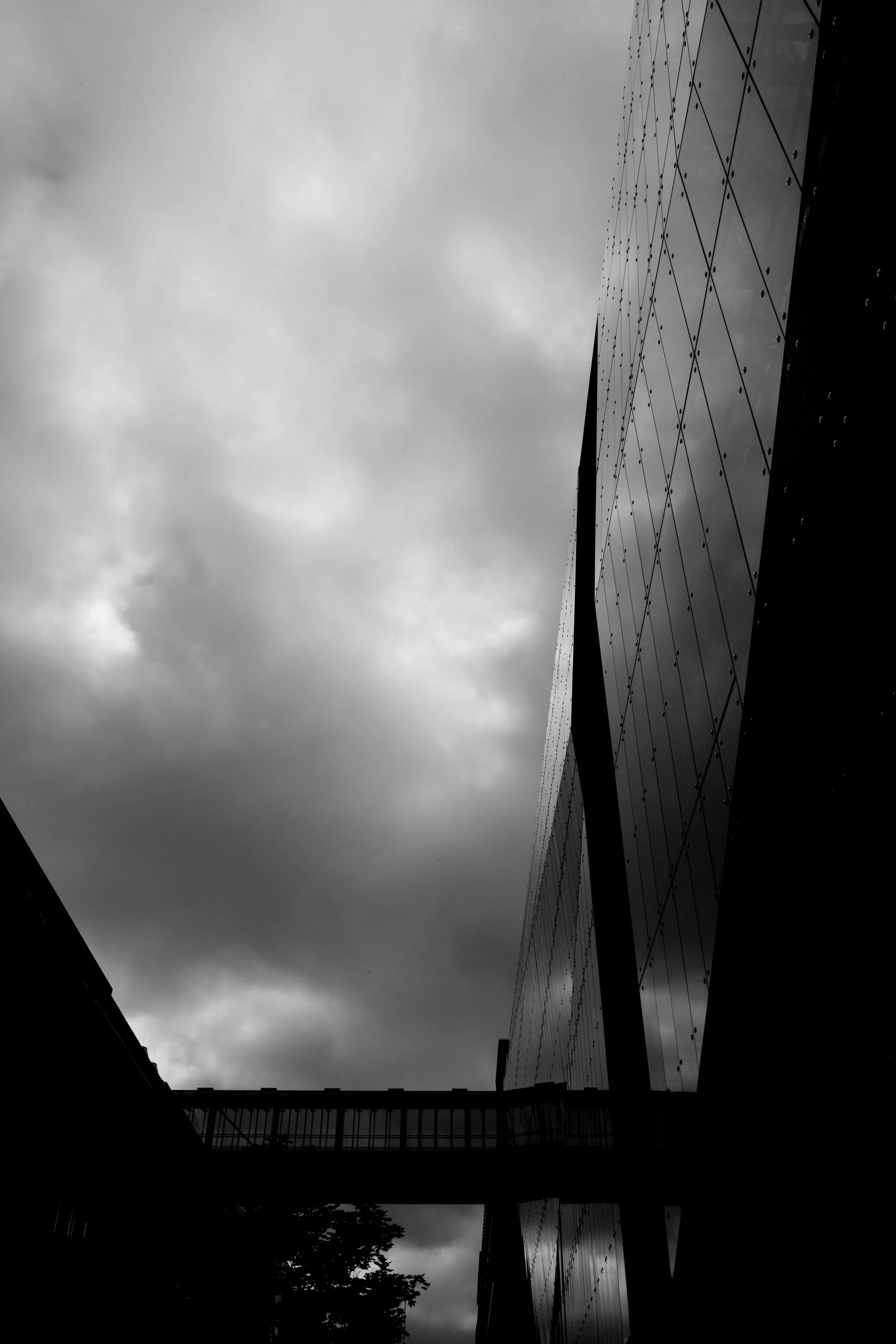 Black and white photo of modern building with reflective glass windows and a bridge connecting two structures, under a cloudy sky.