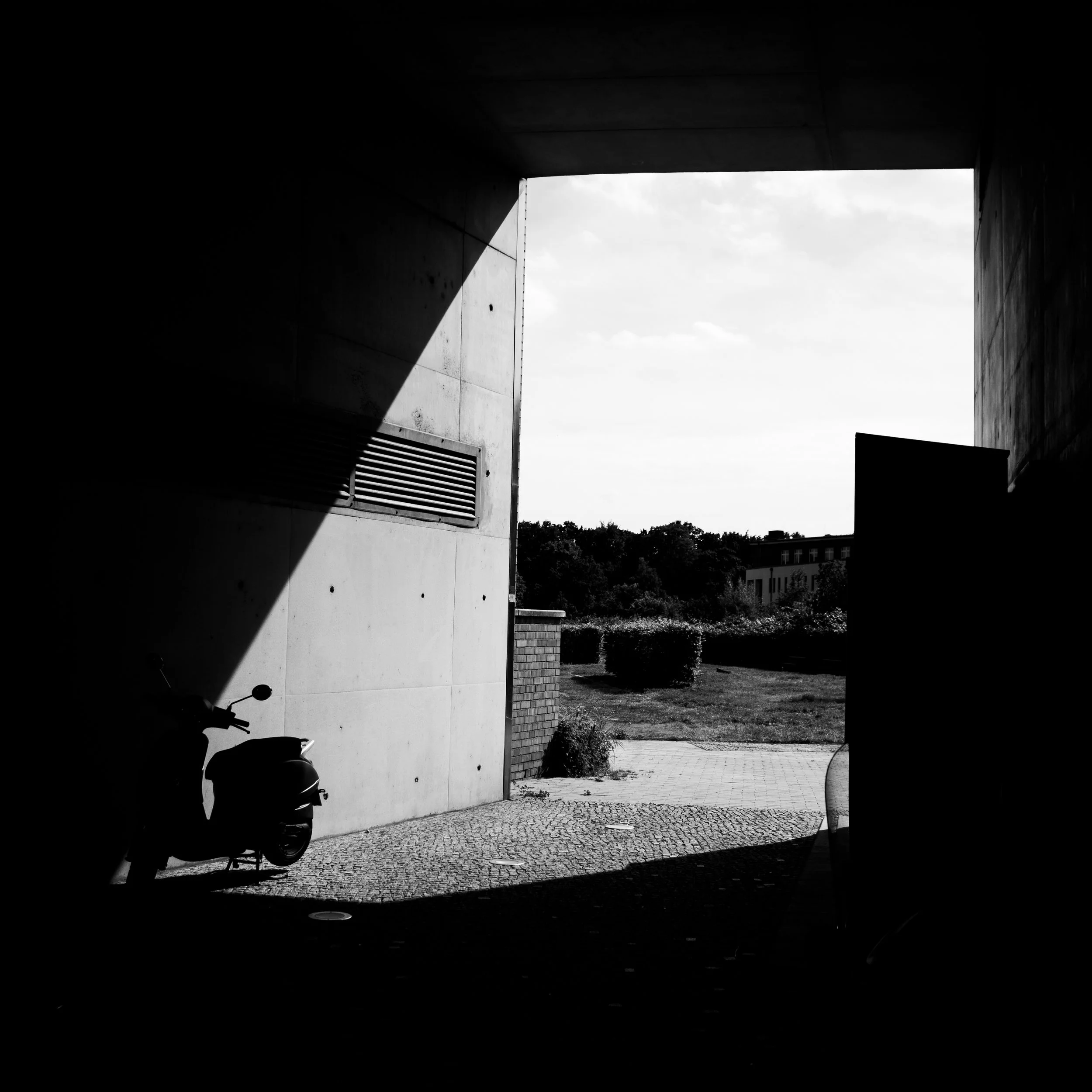 View of an outdoor scene through a dark tunnel, showing a parked scooter, bushes, and a building in the distance.