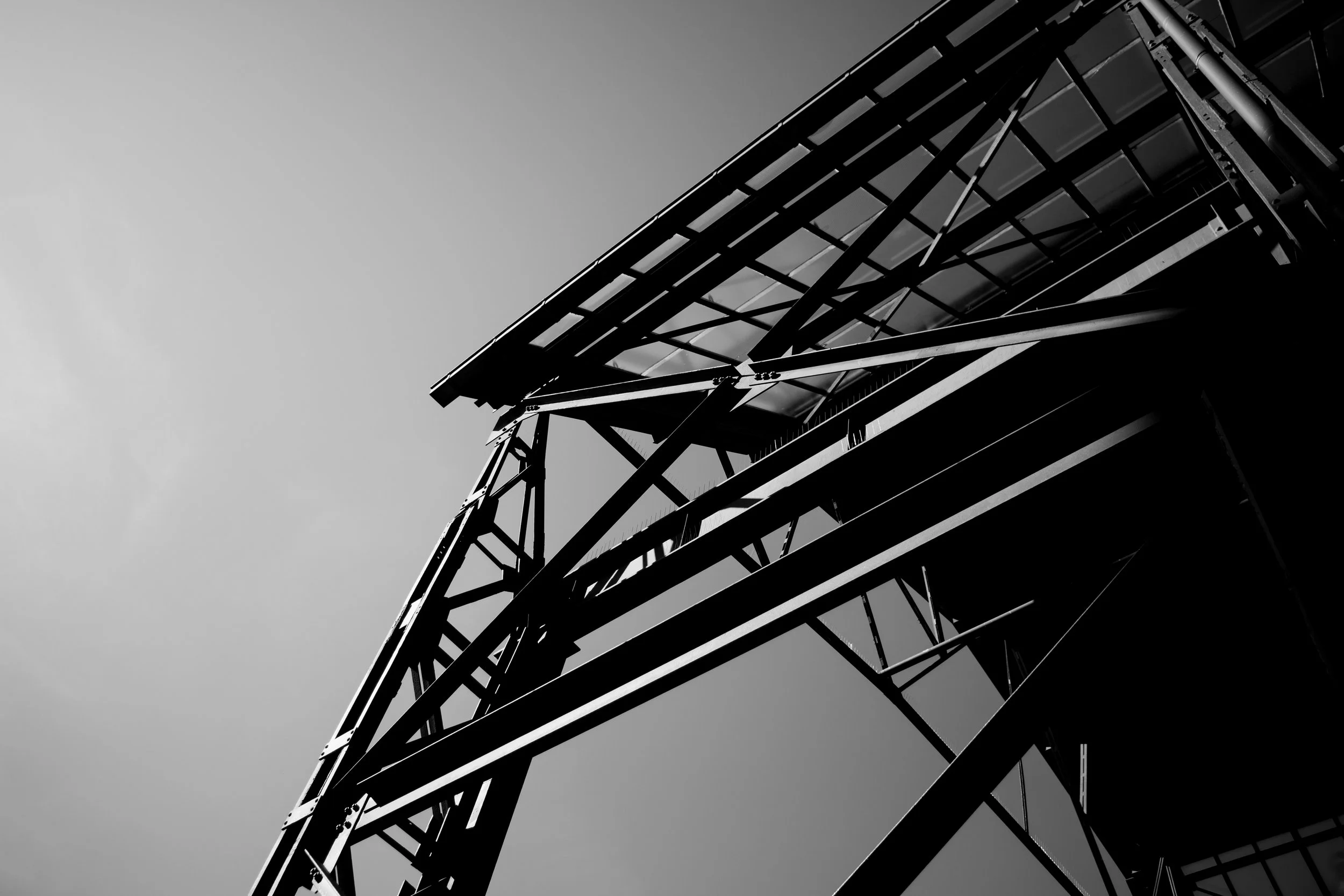 A black and white photo of a tall metal tower with structural beams, viewed from below against a clear sky.
