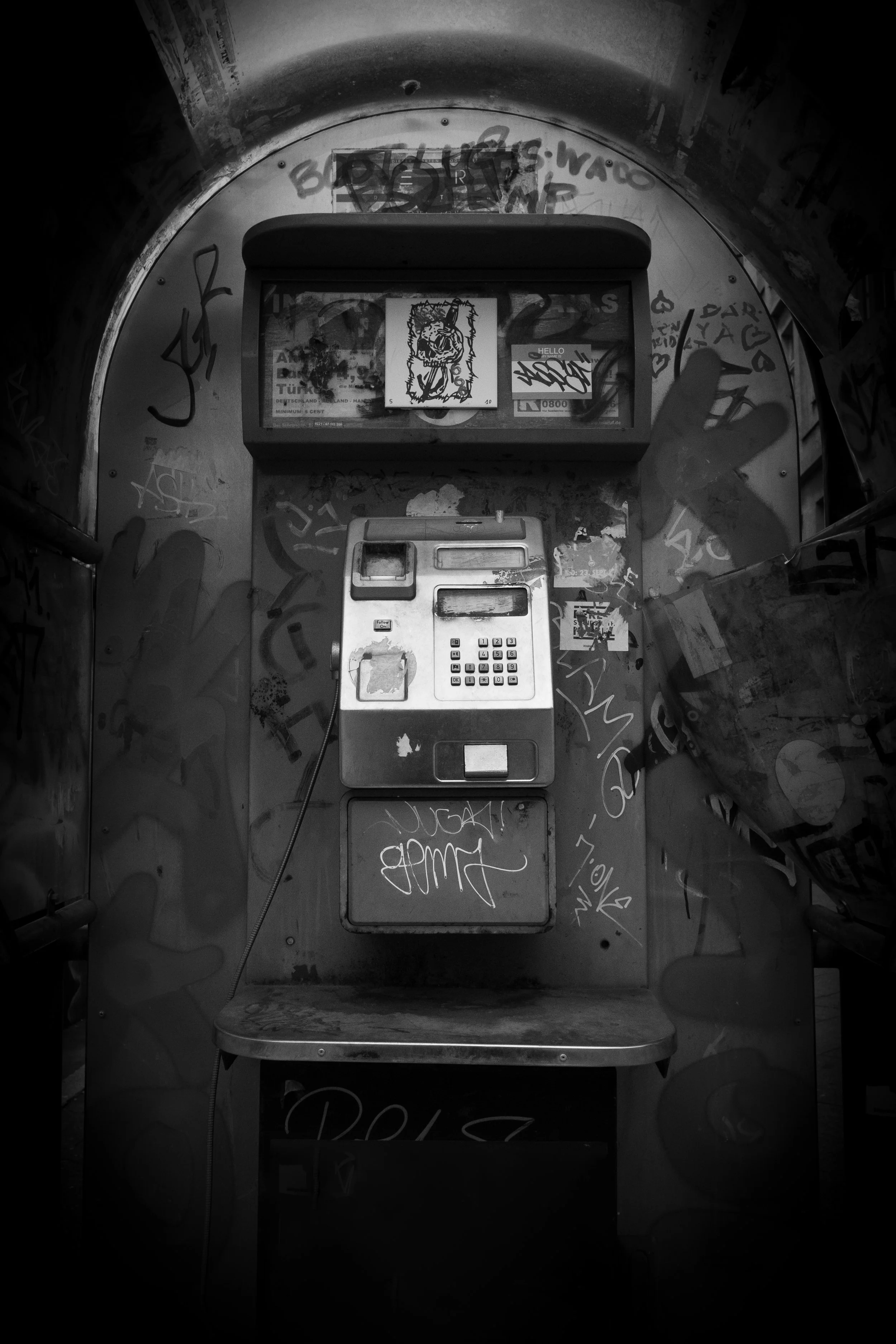 Old public payphone mounted inside a graffiti-covered booth with a scratched display, surrounded by graffiti tags and stickers, in black and white.