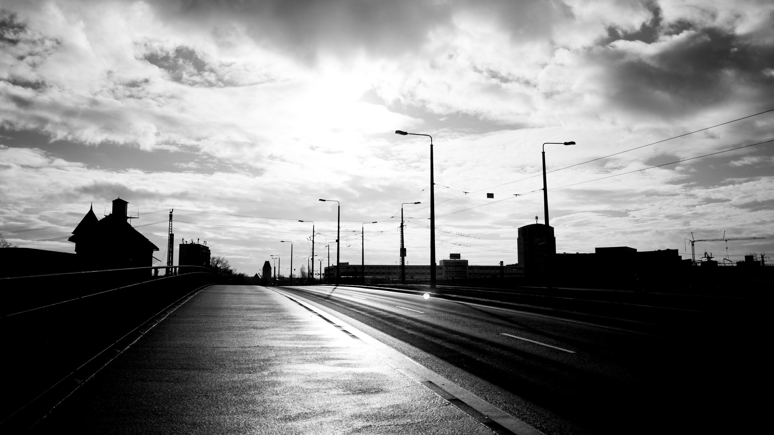 Black and white photo of an empty city street with streetlights and buildings in the distance under a cloudy sky with the sun shining through.