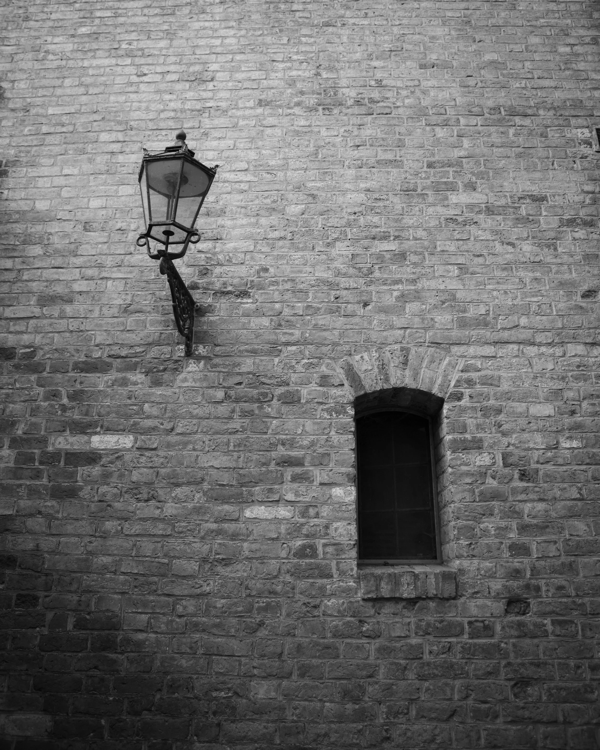 A black and white photo of a brick wall with a vintage street lamp mounted on the wall and a small window with a stone arch above it.